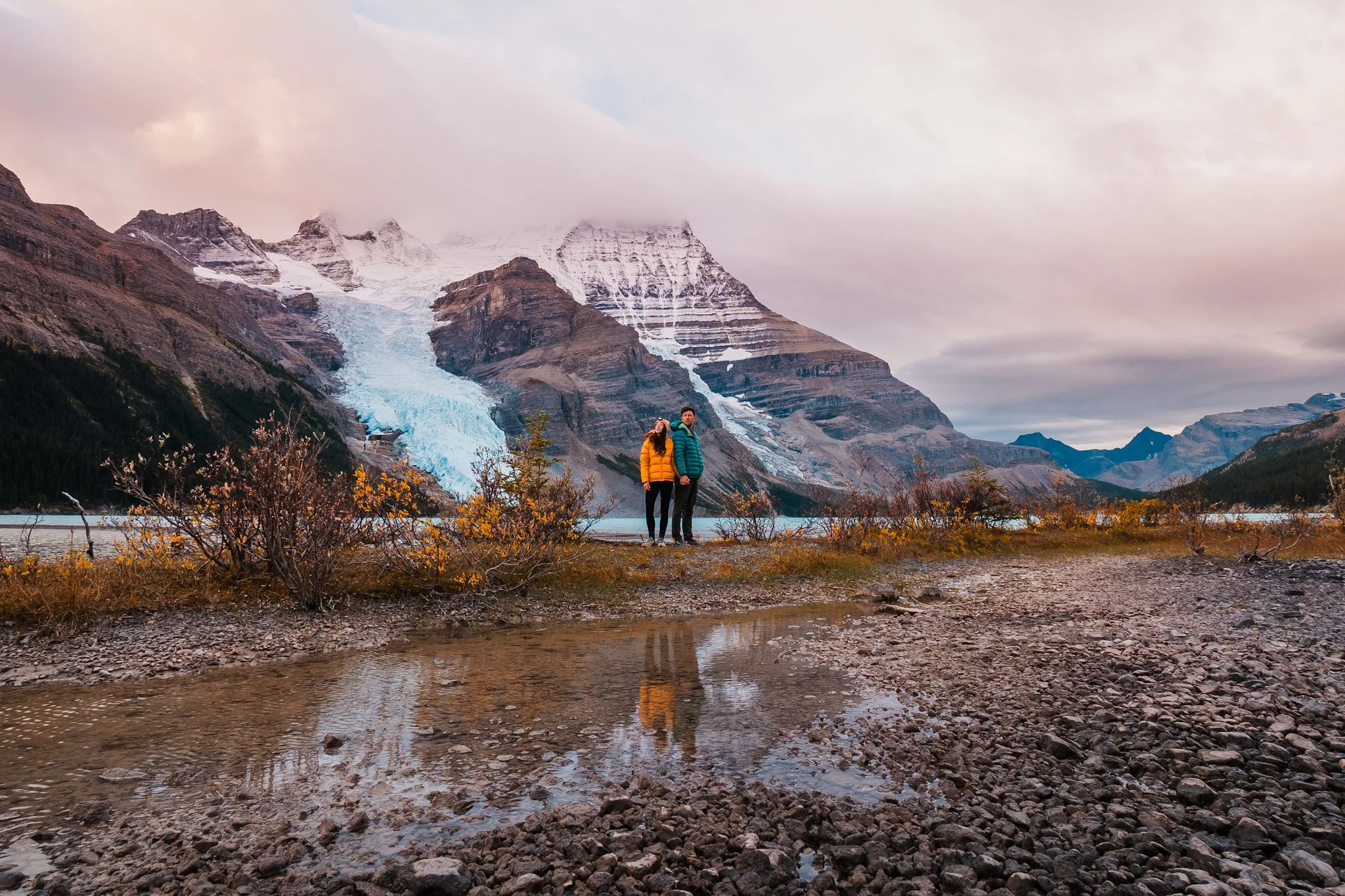 a man and women standing together in down jackets and hiking clothes, in front of a big mountain with a glacier and alpine lake