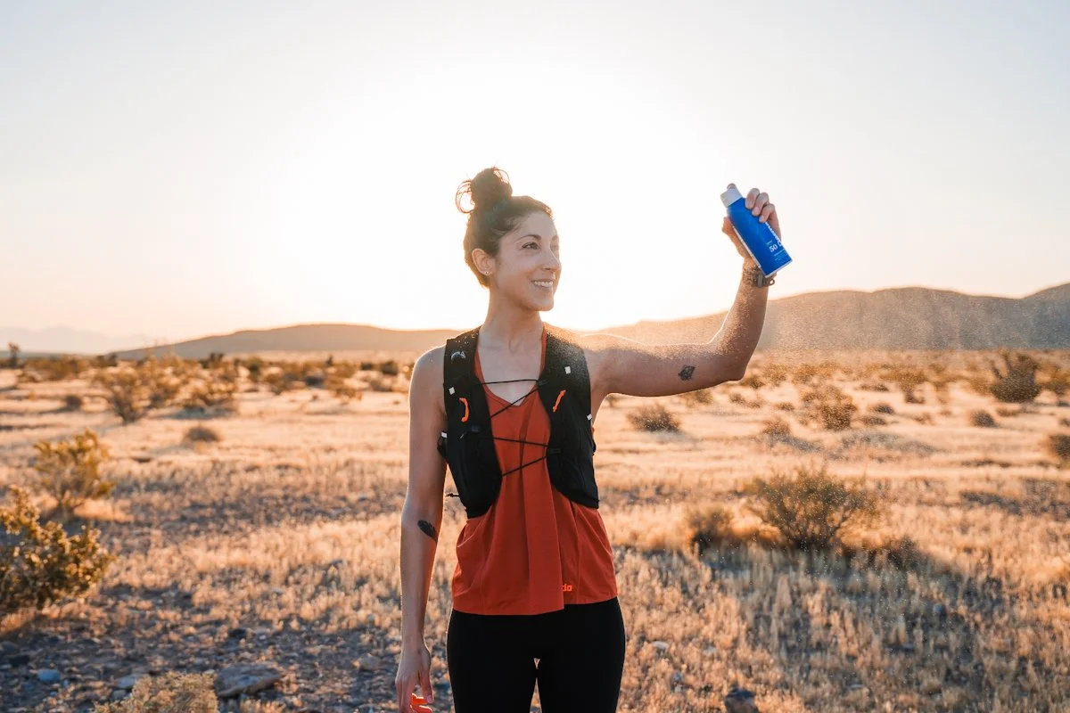 Woman hiking in a desert landscape at sunset, holding a blue water bottle and smiling.