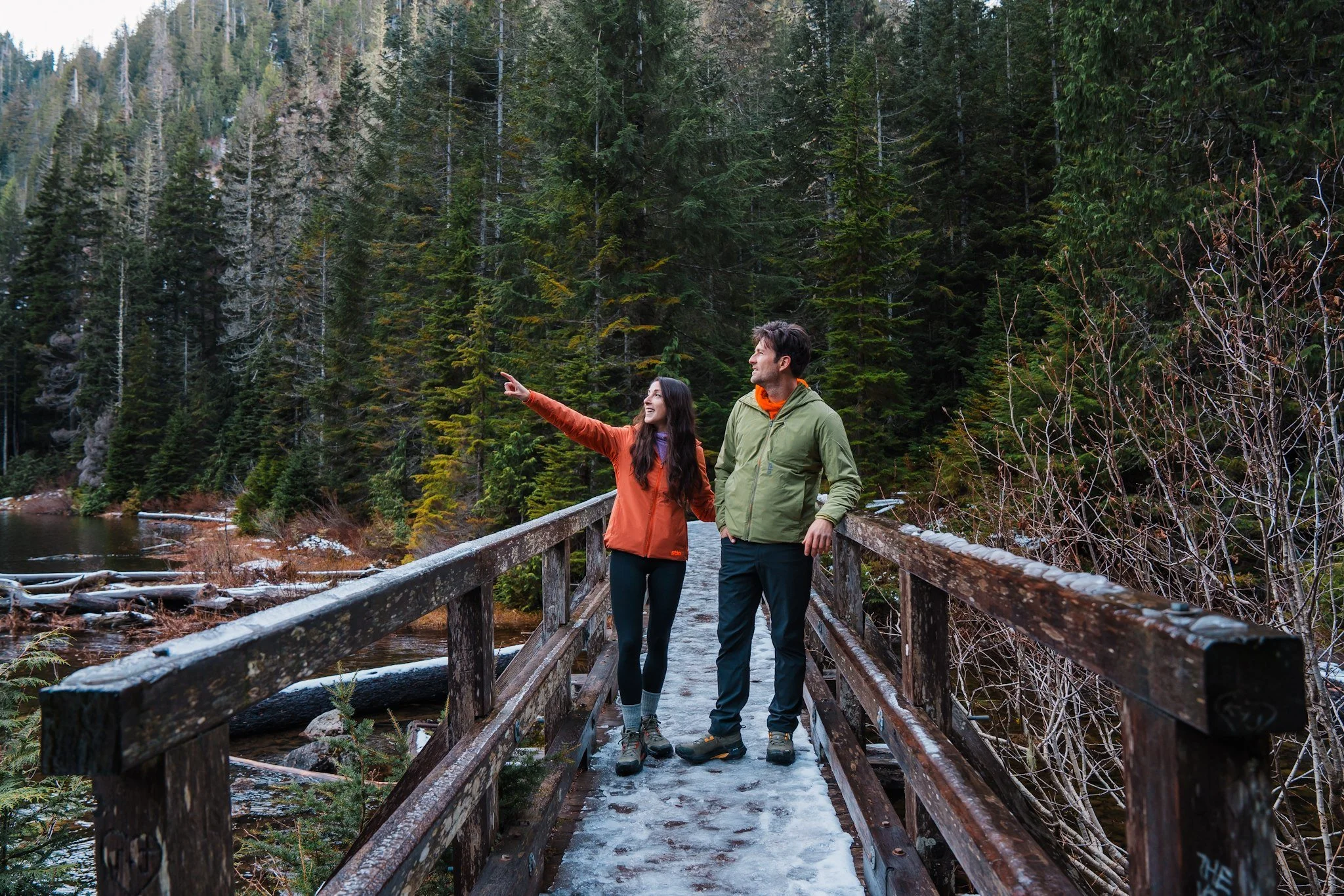 A couple standing on a snowy bridge while hiking, the woman is pointing and wearing an orange jacket and black leggings, the man is wearing a light green jacket and black hiking pants