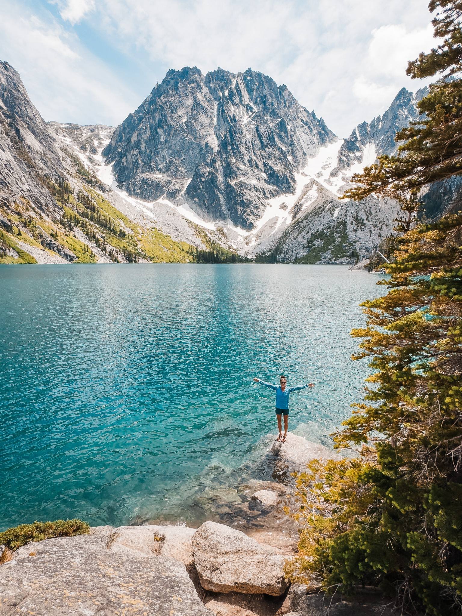 a hiker standing on a rock next to a bight blue alpine lake with rocky peaks in the background