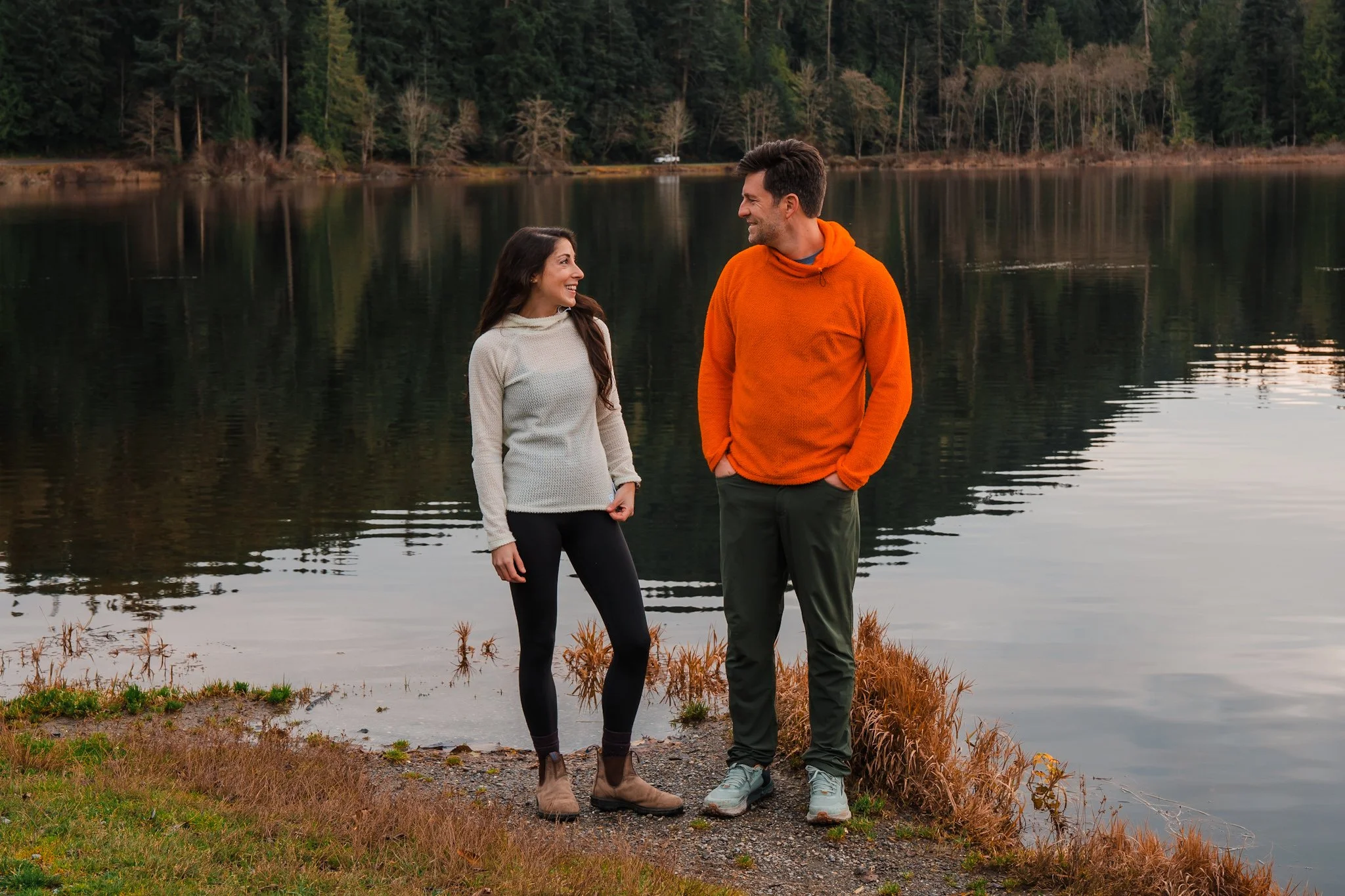 a couple standing in front of a lake, the woman is wearing a cream colored fleece hoodie and the man is wearing an orange fleece