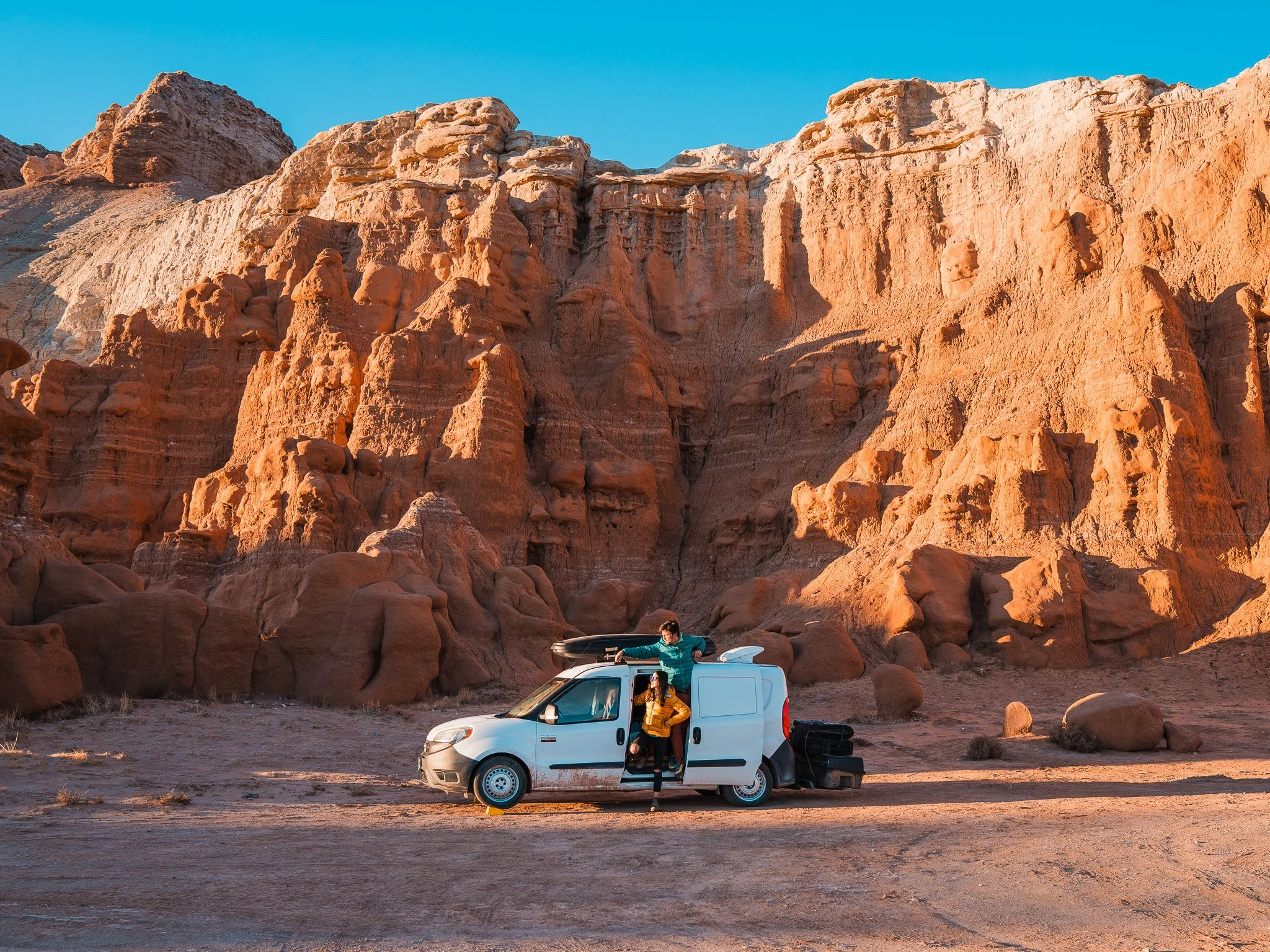 a couple with their small, white van, parked in the desert with a red sandstone wall behind them
