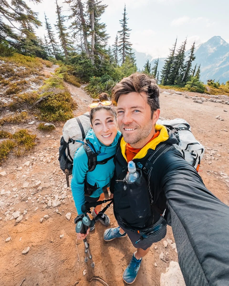 a couple taking a selfie, smiling for the camera with backpacks on while backpacking in the North Cascades