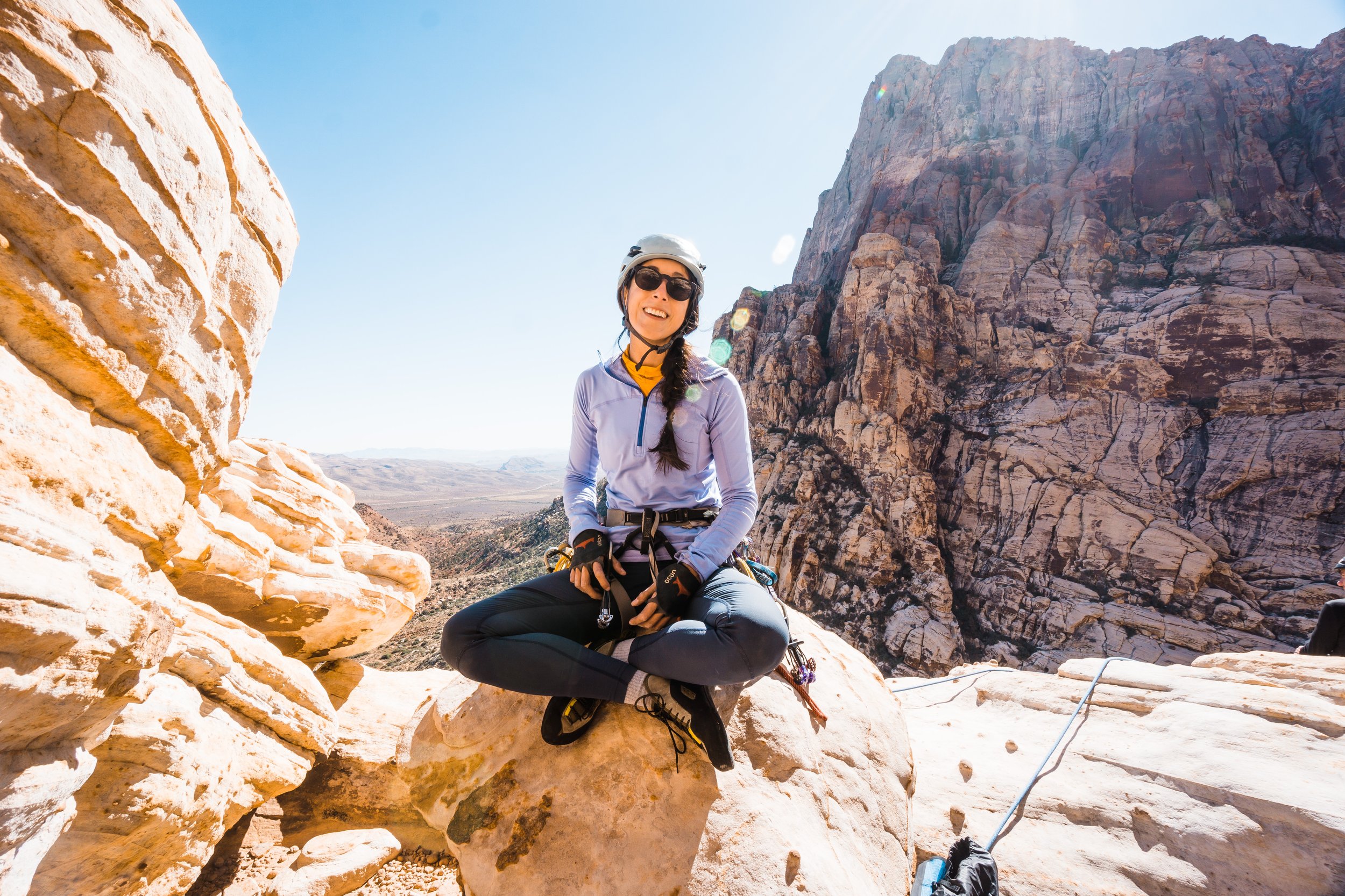 A woman in climbing gear sitting on a rock with a desert landscape, cliffs, and blue sky in the background.