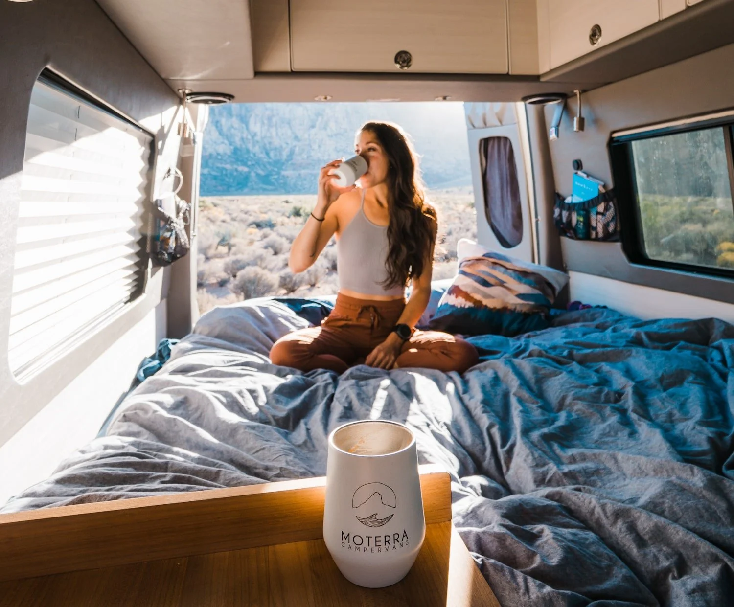 A woman on the bed, inside of a campervan, drinking coffee with the back doors open.