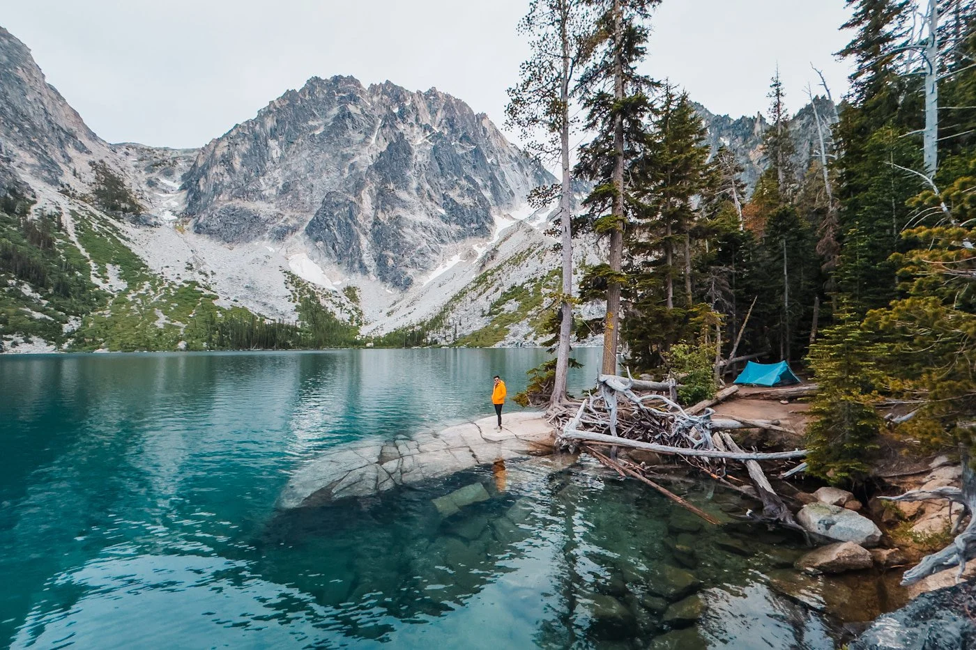 a man standing on a rock next to colchuck lake with his tent on one side and dragonatil peak to the other