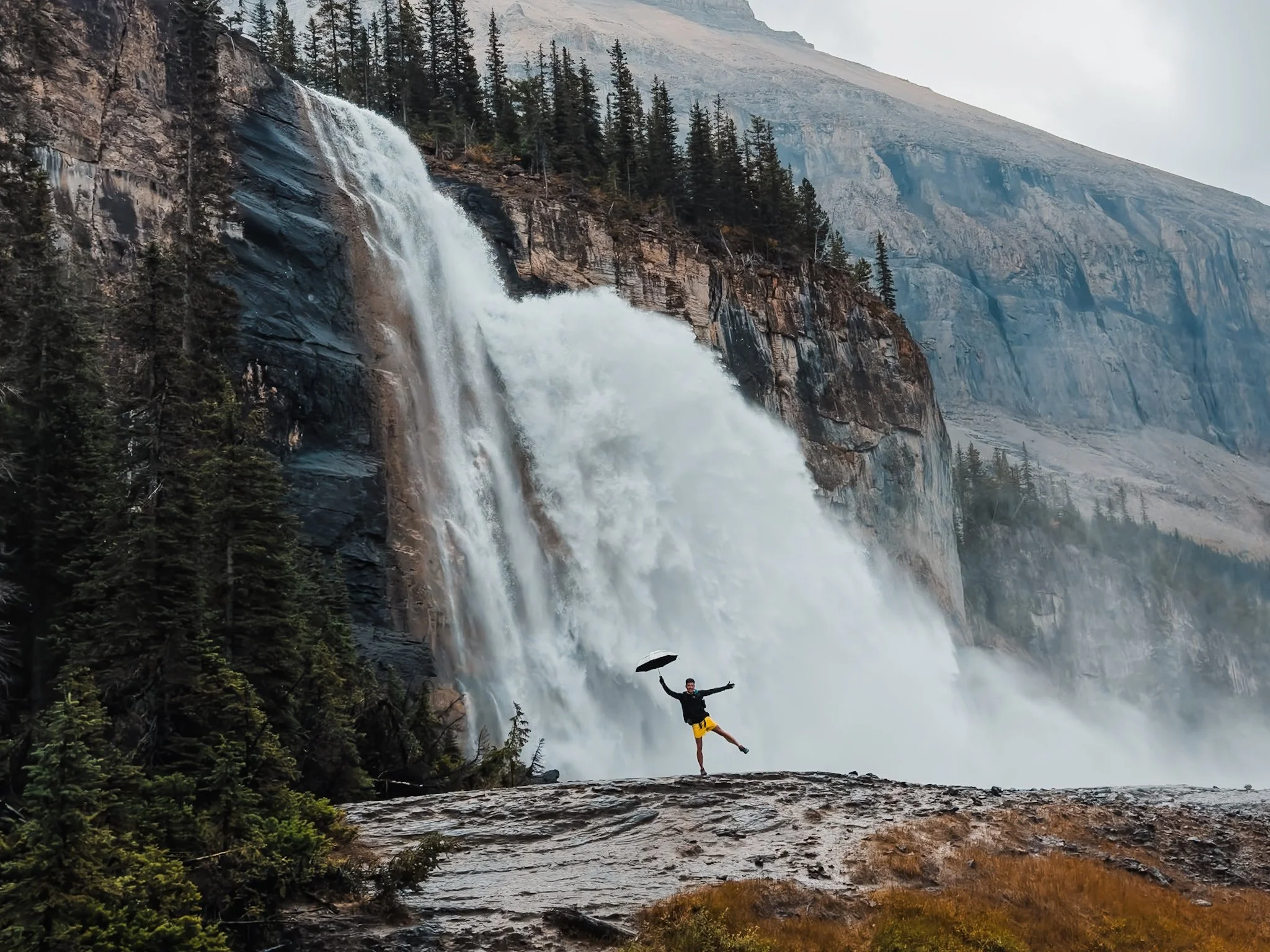 a small person holding an umbrella, standing in front of a huge waterfall in the mountains