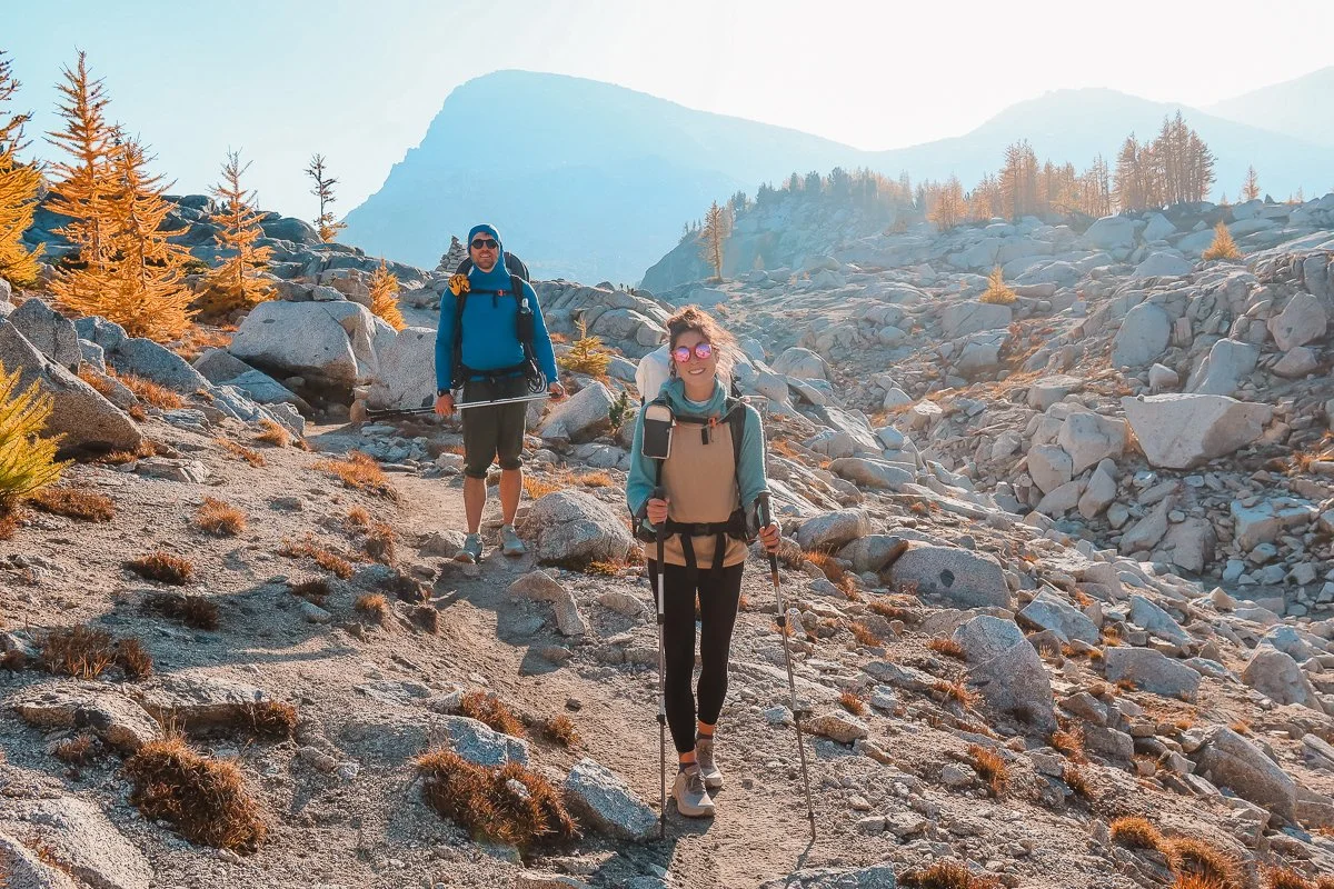 Two backpackers hiking on trail in the core enchantments with some mountains and larch trees in the background
