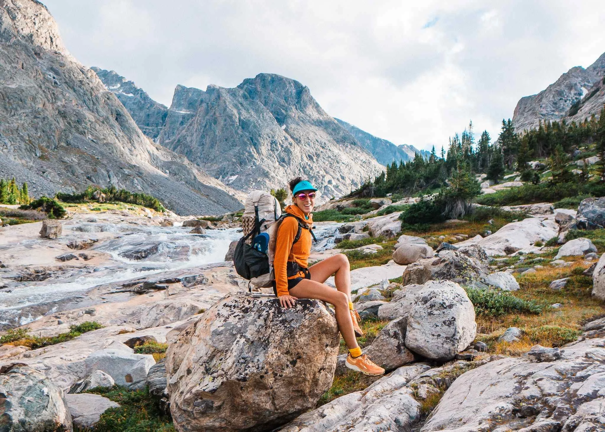 A woman with a blue visor, orange jacket, black shorts, and orange shoes sitting on a large rock in a mountainous landscape with a river, rocks, trees, and towering mountains in the background.