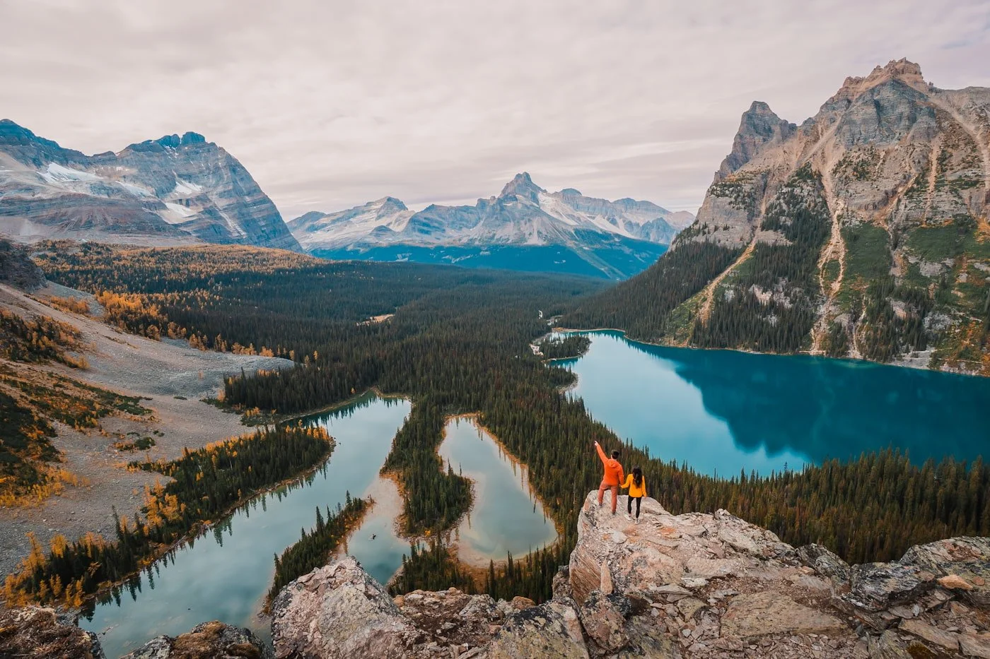 a couple standing at an overlook above Lake O'Hara with lakes below and mountains in the distance