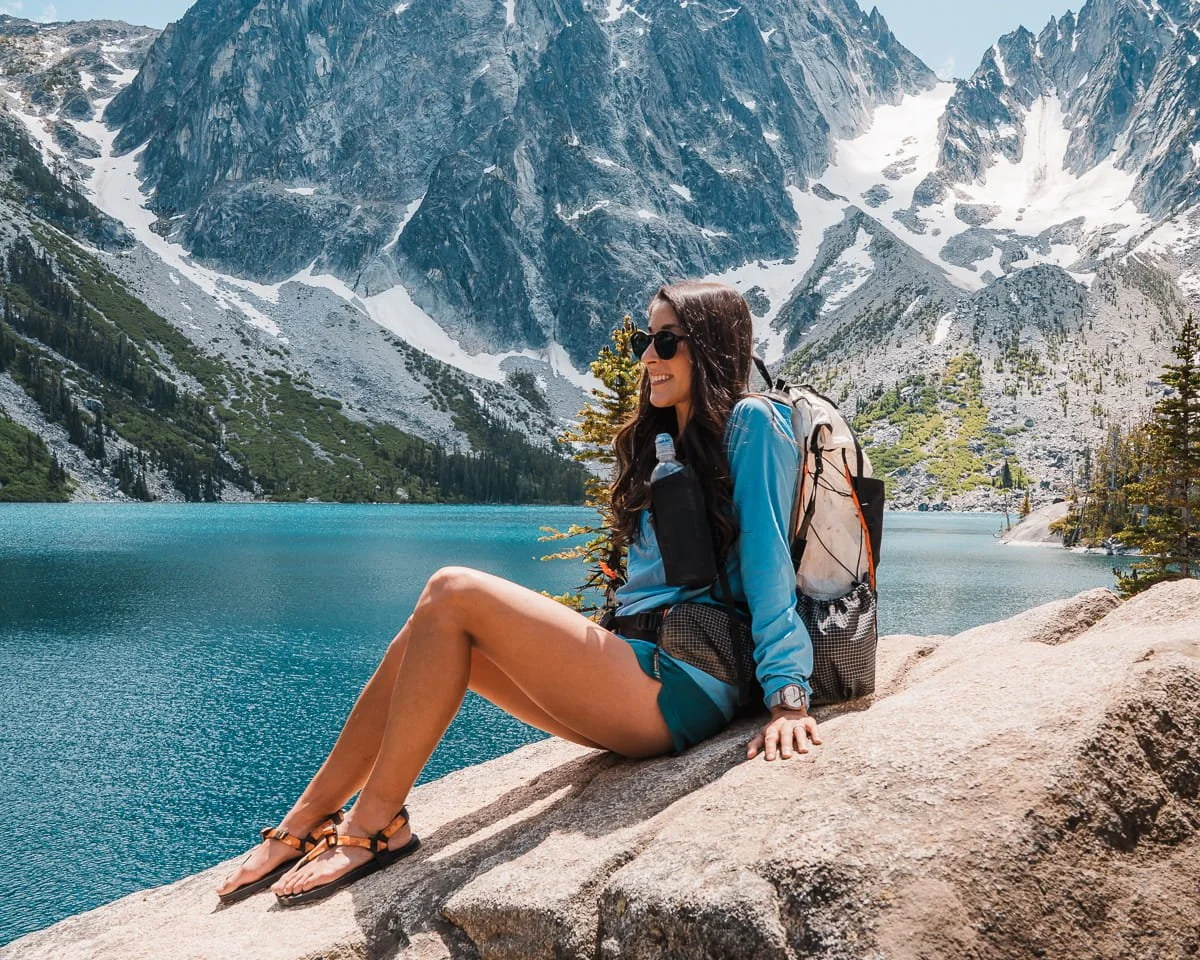 a woman sitting on a rock in front of a bright blue lake with a blue sun hoodie and backpack on