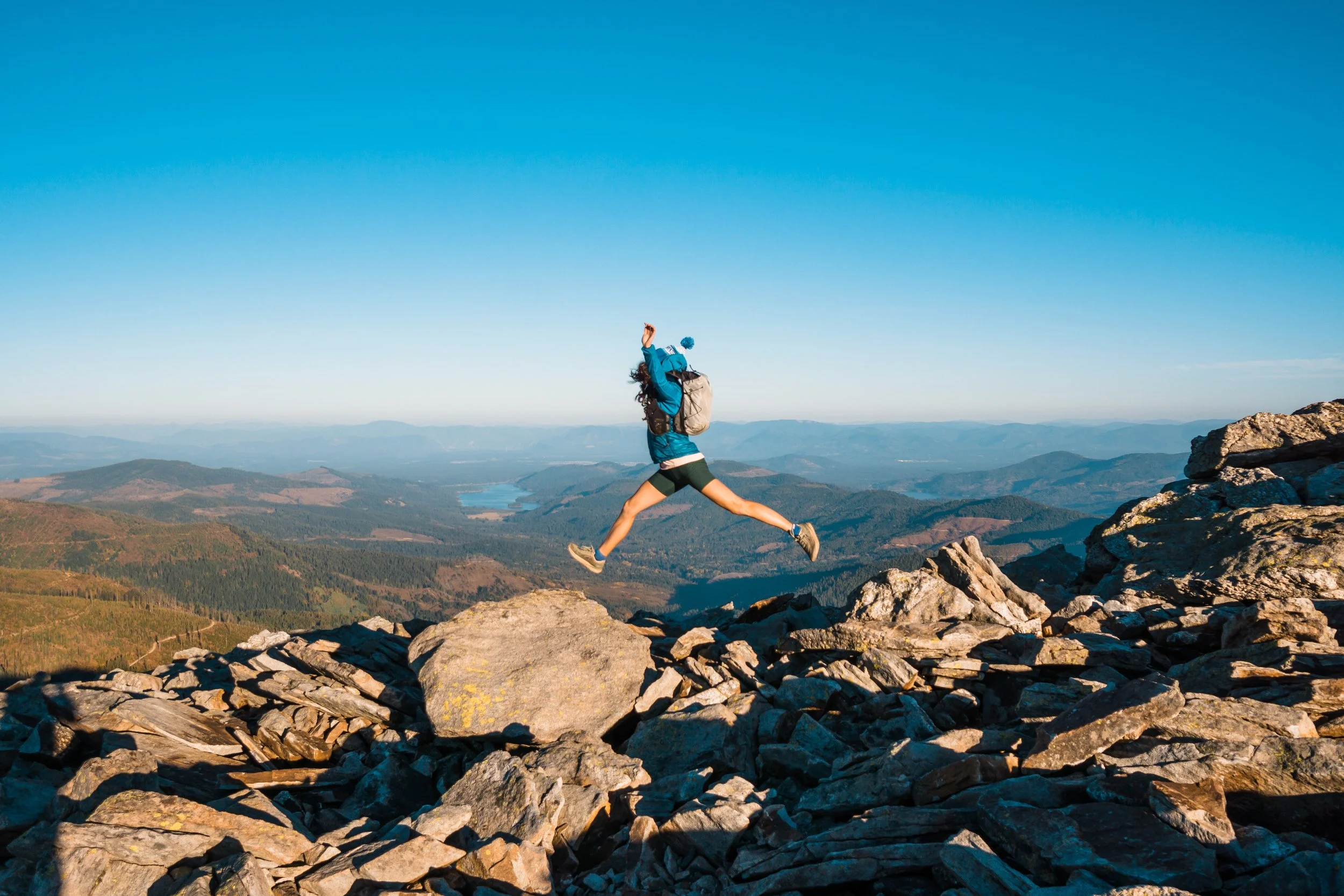 A person wearing outdoor hiking gear jumping over rocks on a mountain summit during daytime, with rolling hills and a lake in the distance under a clear blue sky.