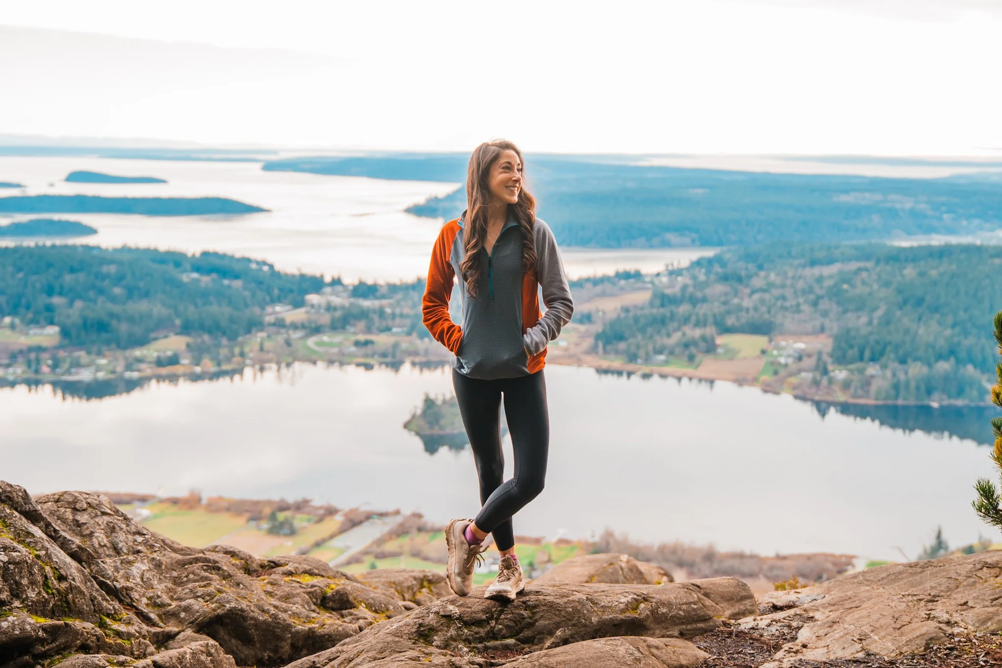 a woman standing at a view point with lakes and the sound in the background. She is wearing a grey and burnt orange fleece pullover