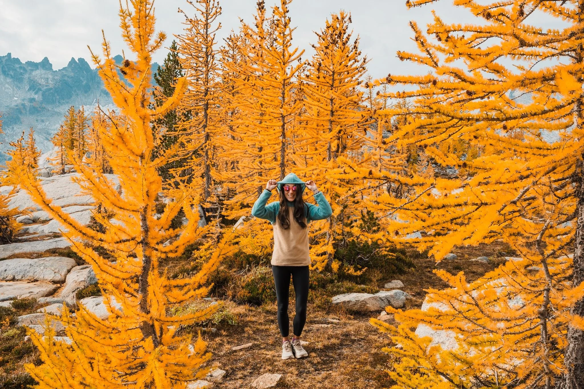 a woman standing amongst golden larch trees, wearing a turquoise and brown fleece hoodie with the hood up