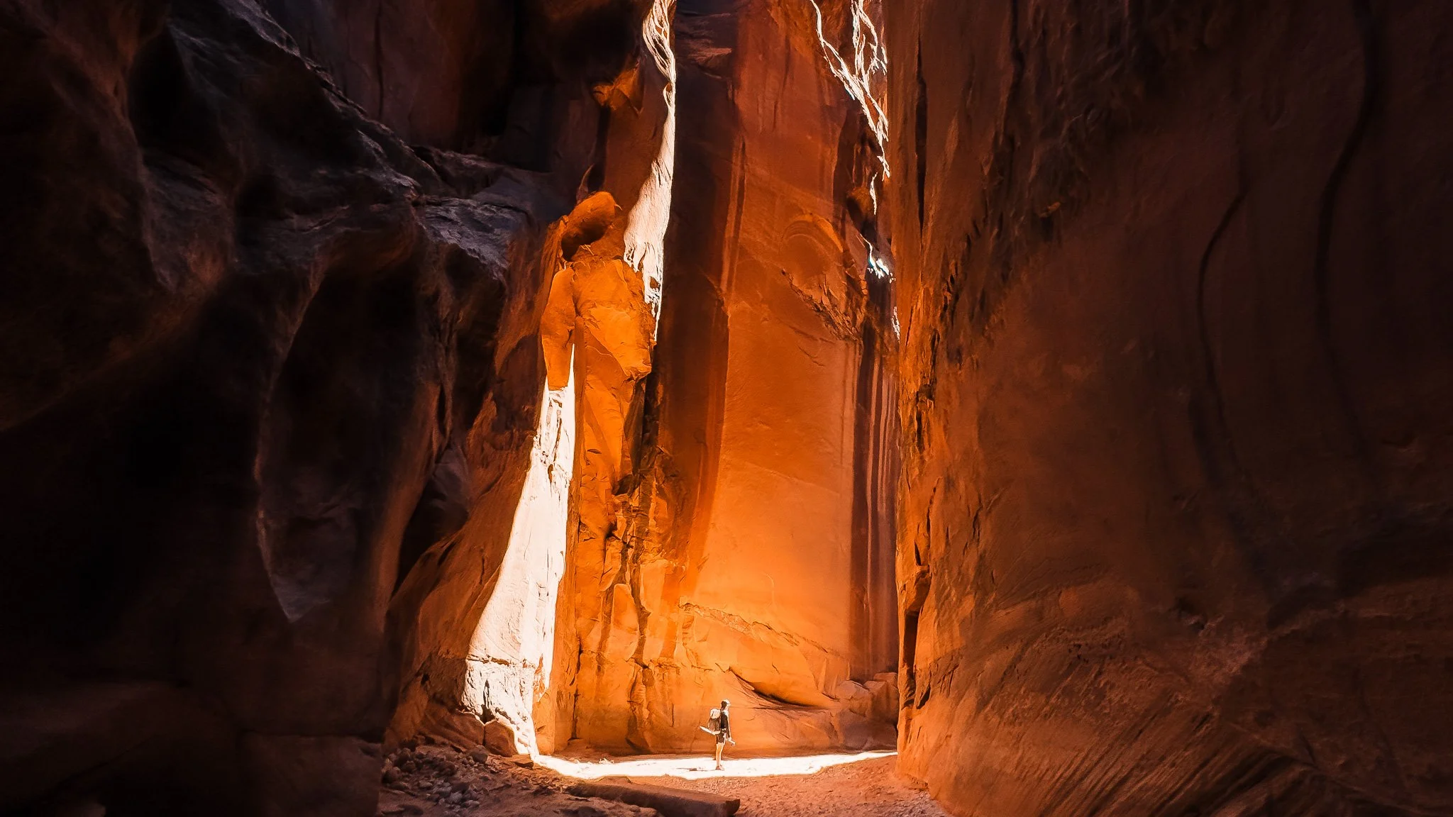a tiny backpacker hiking through a slot canyon with high sandstone walls and a section illuminated by sunlight streaming through