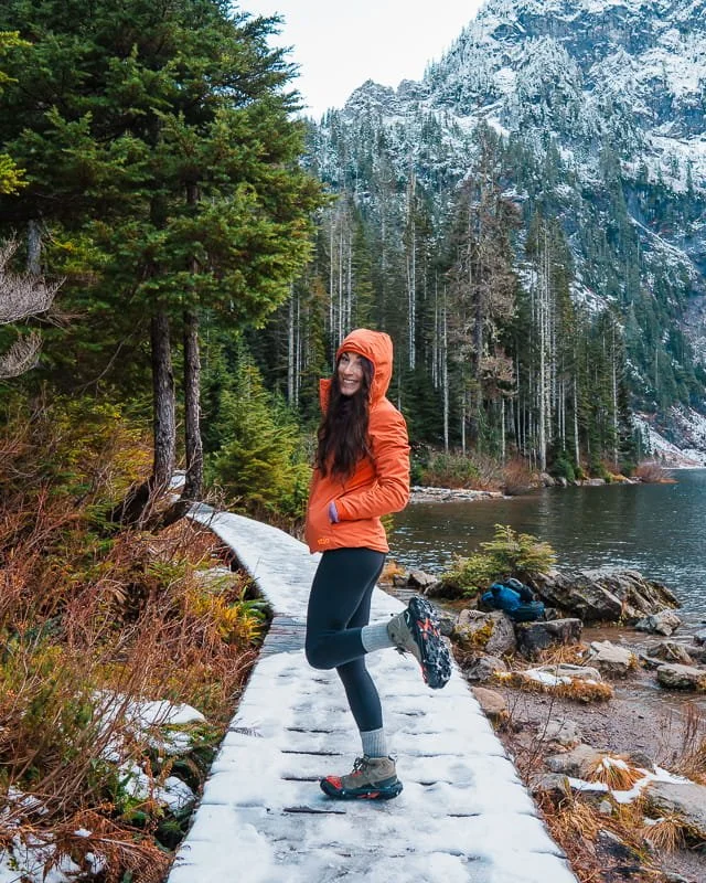 a woman hiking on a snowy board walk next to an alpine lake, wearing an orange puffy jacket, black leggings, hiking boots, and microspikes