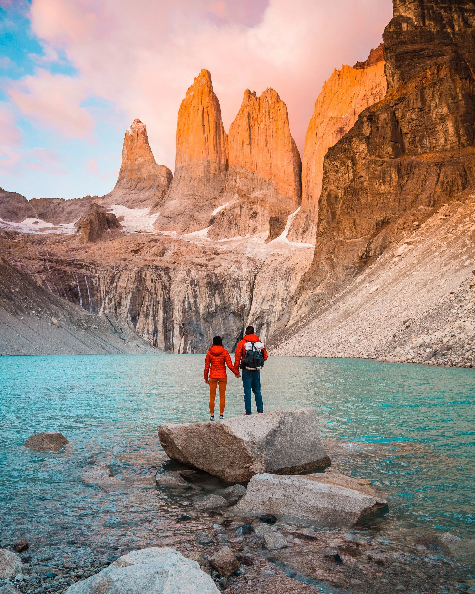 a couple in red, standing on a rock in front of a turquoise lake and the sun rising on towering mountains