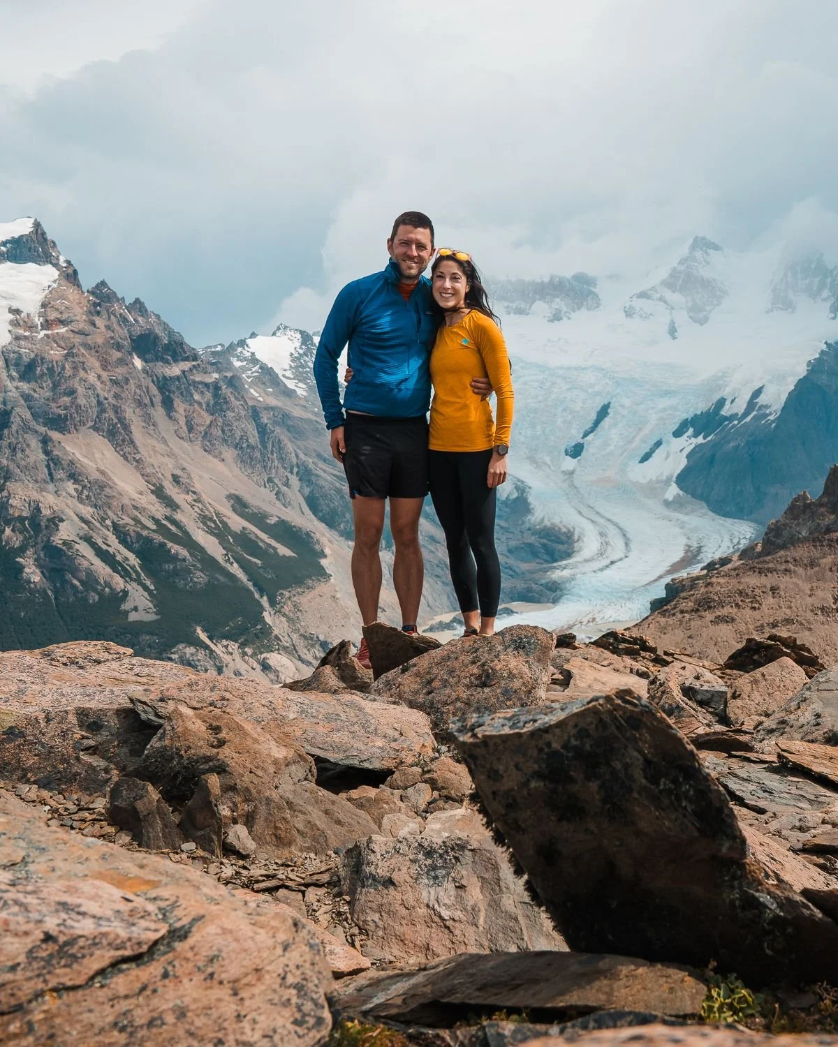 Two hikers, a man and a woman, standing close together on rocky terrain with a mountain glacier in the background, wearing outdoor gear.