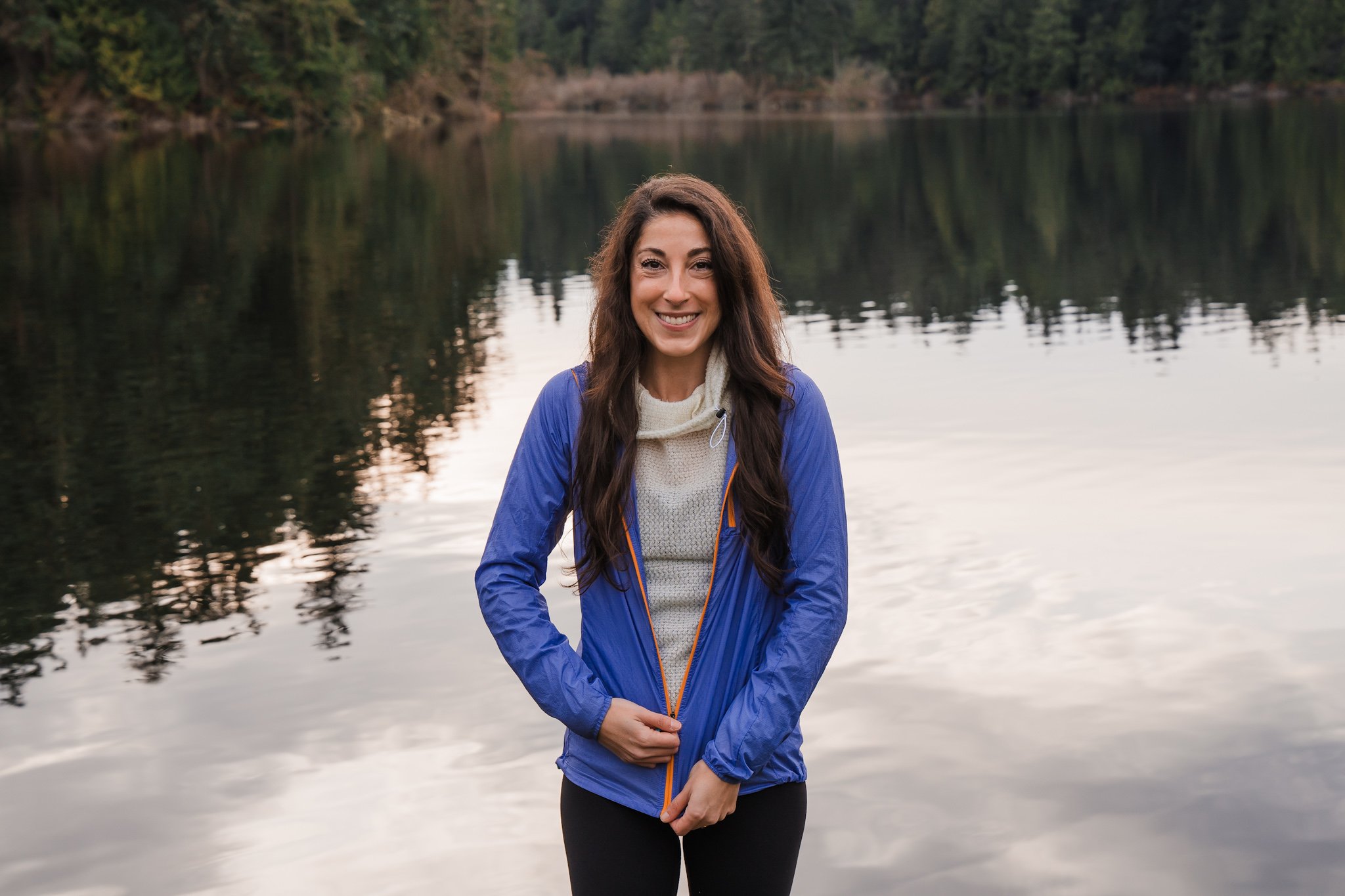 A woman standing in front of a lake, wearing a white alpha direct fleece and zipping up a purple wind jacket over.