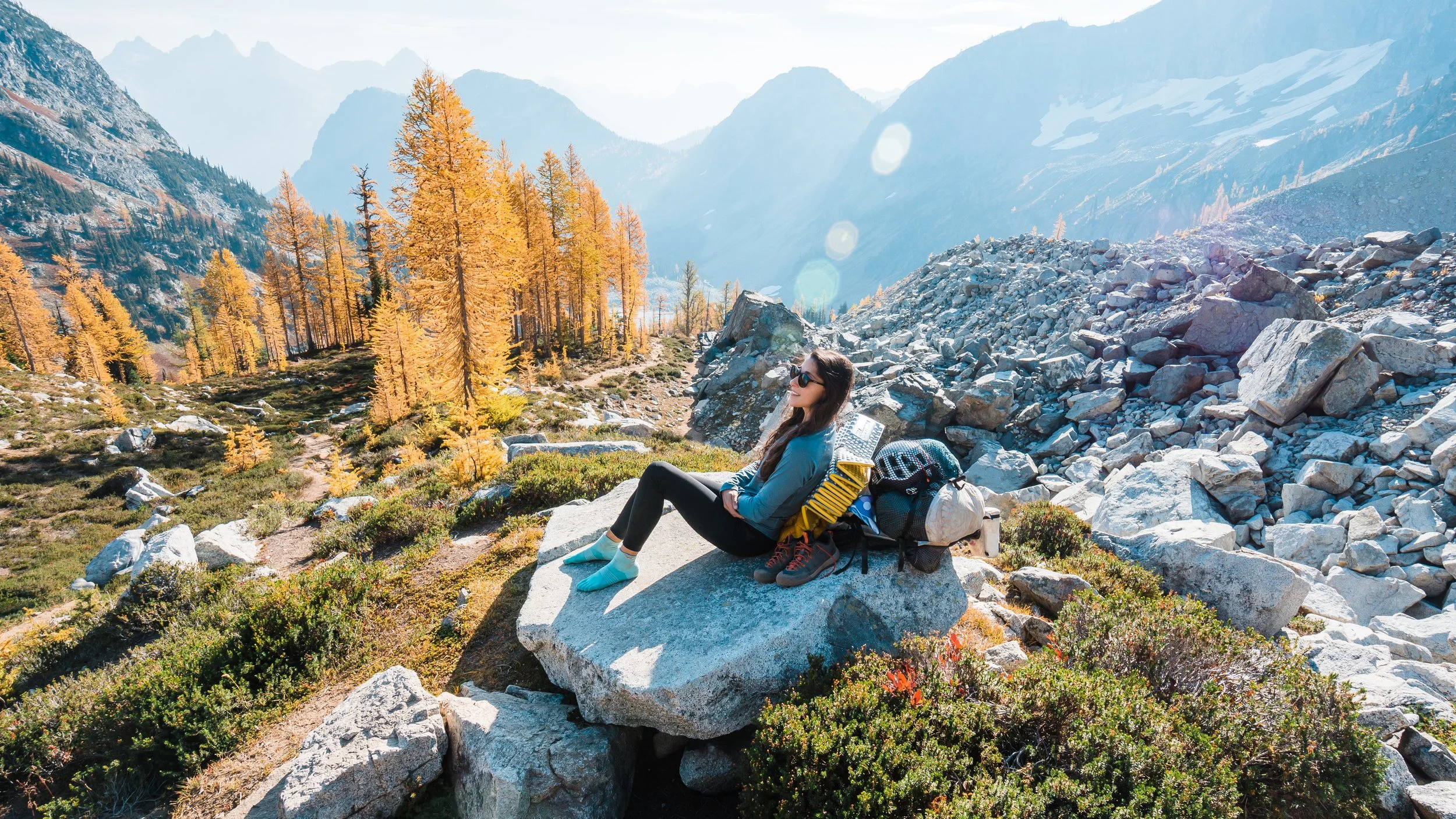 A woman with sunglasses resting on a large rock in a mountainous landscape during fall, with orange trees, rocky terrain, and snow-capped peaks in the background.