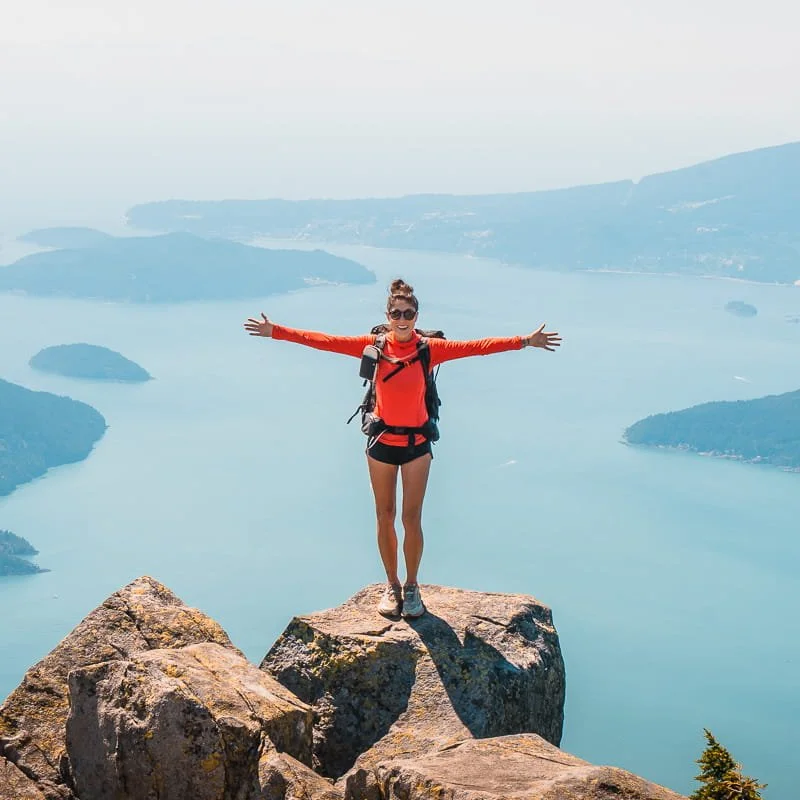 a woman backpacker standing on. a rock with her arms outstretched, wearing a bright pink sun hoodie, in front of the Howe Sound in British Columbia