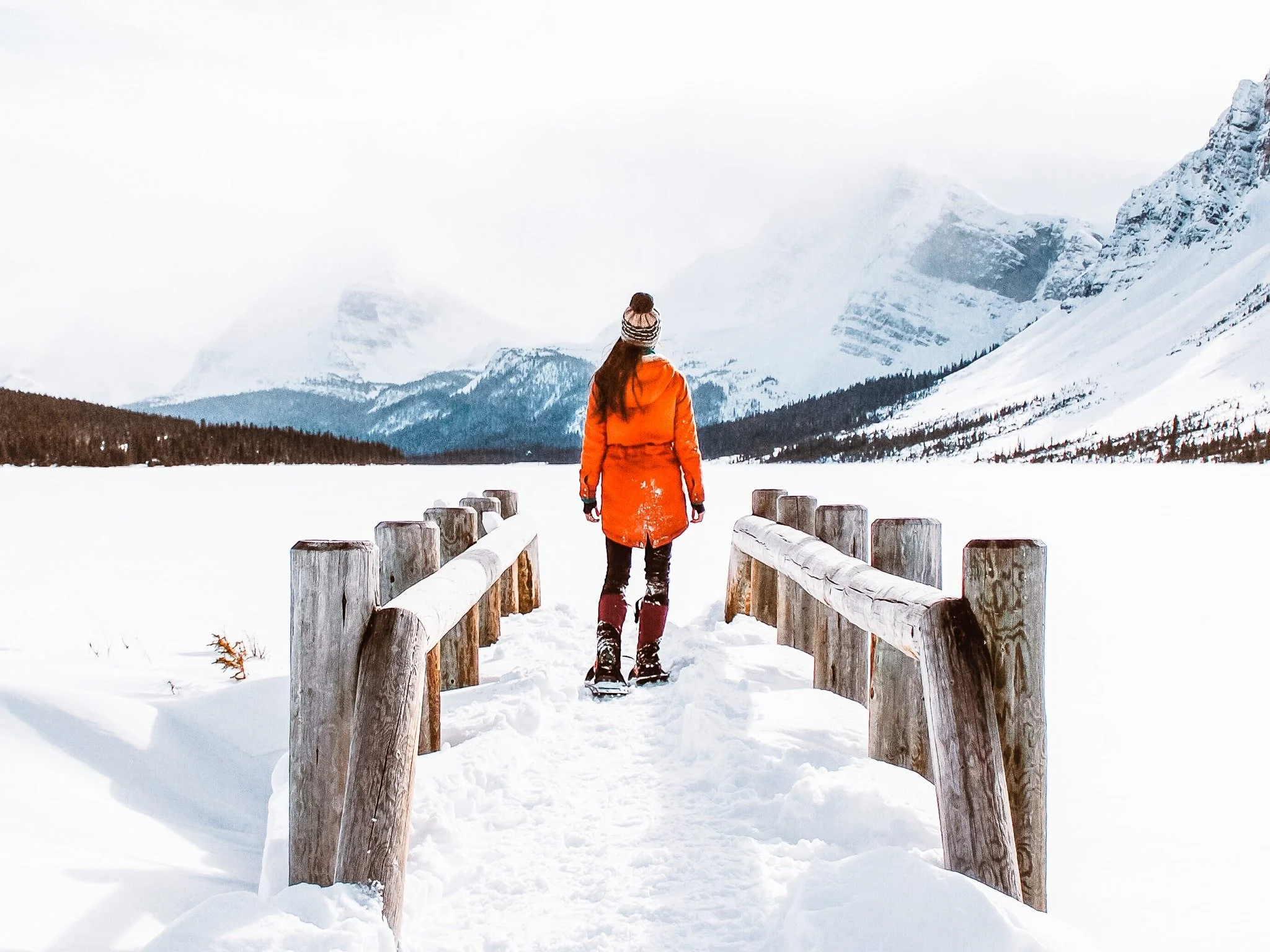 a woman wearing a bright orange jacket and snowhoes, walking across the bridge at Bow lake in Banff, with the lake covered in snow and snowy mountains in the background