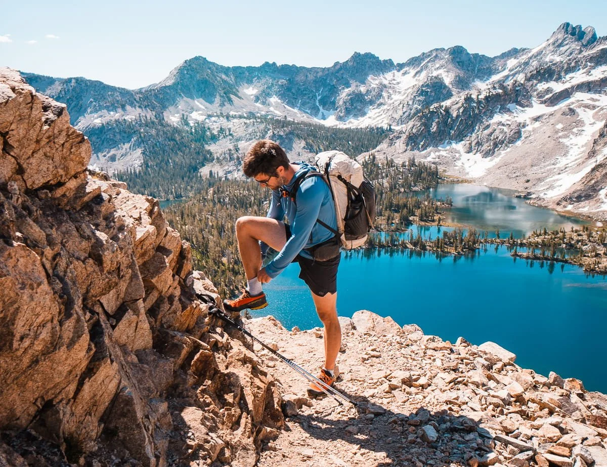a man backpacking, wearing a blue sun shirt and shorts, pulling up his sock. With a bright lue lake and mountains in the background