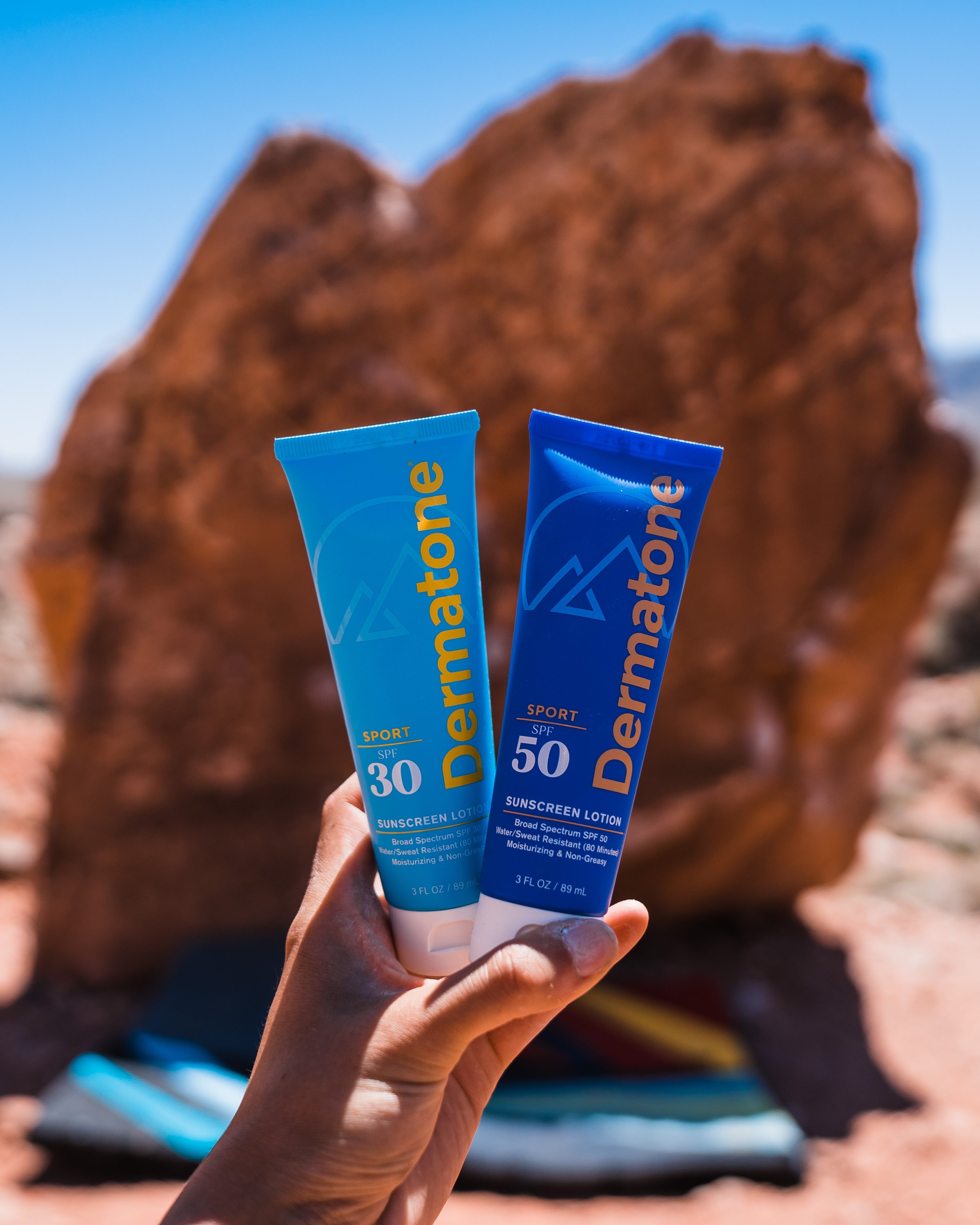 A hand holding two tubes of Dermatone sunscreen lotion, one with SPF 30 and the other with SPF 50, against a rocky outdoor landscape with a large reddish rock in the background and a cloudy blue sky.
