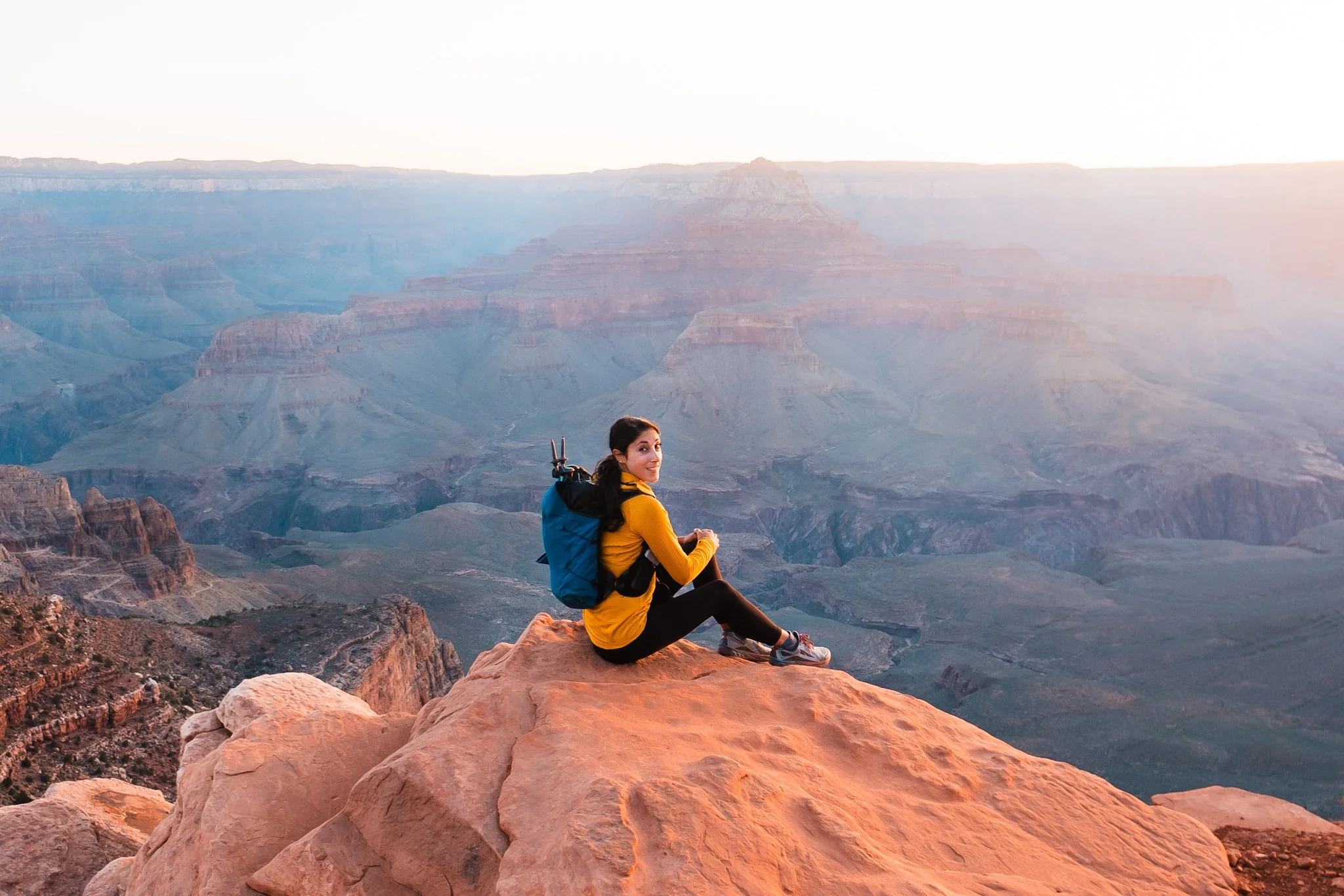 A woman sitting on a rock overlooking the grand canyon at sunrise. She is wearing a yellow fleece and blue trail running backpack