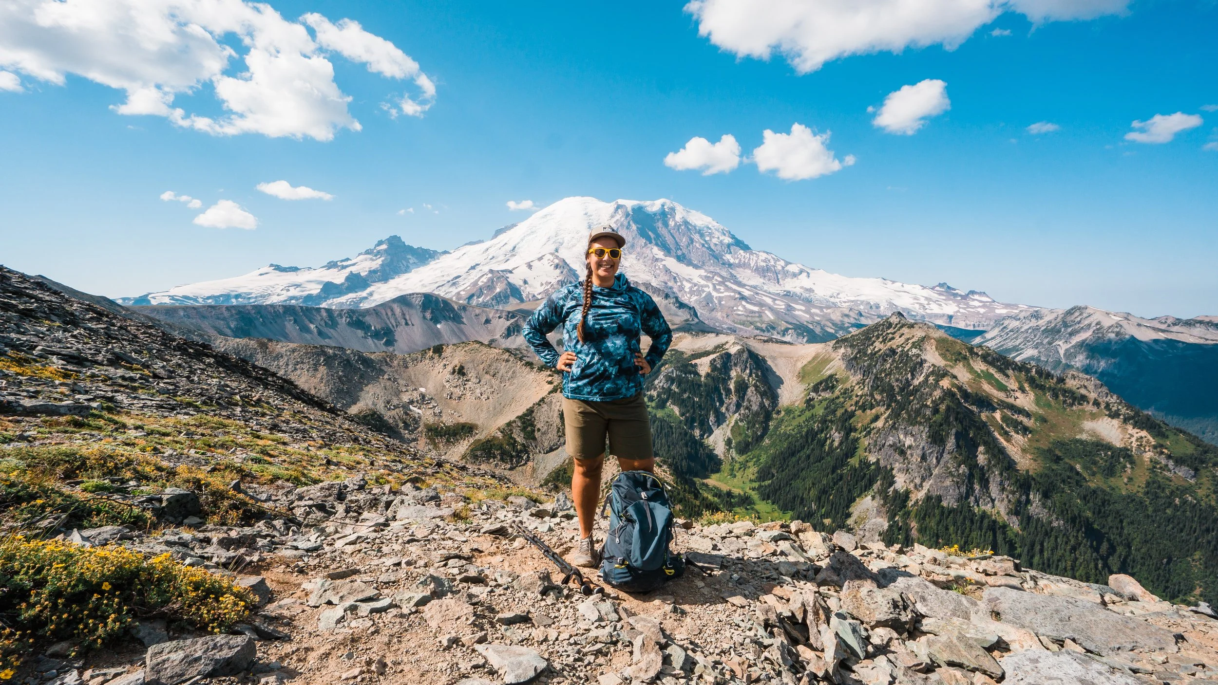 A woman standing on rocky terrain with a mountain and snow-capped peak in the background, wearing sunglasses, a blue patterned jacket, shorts, and hiking shoes.