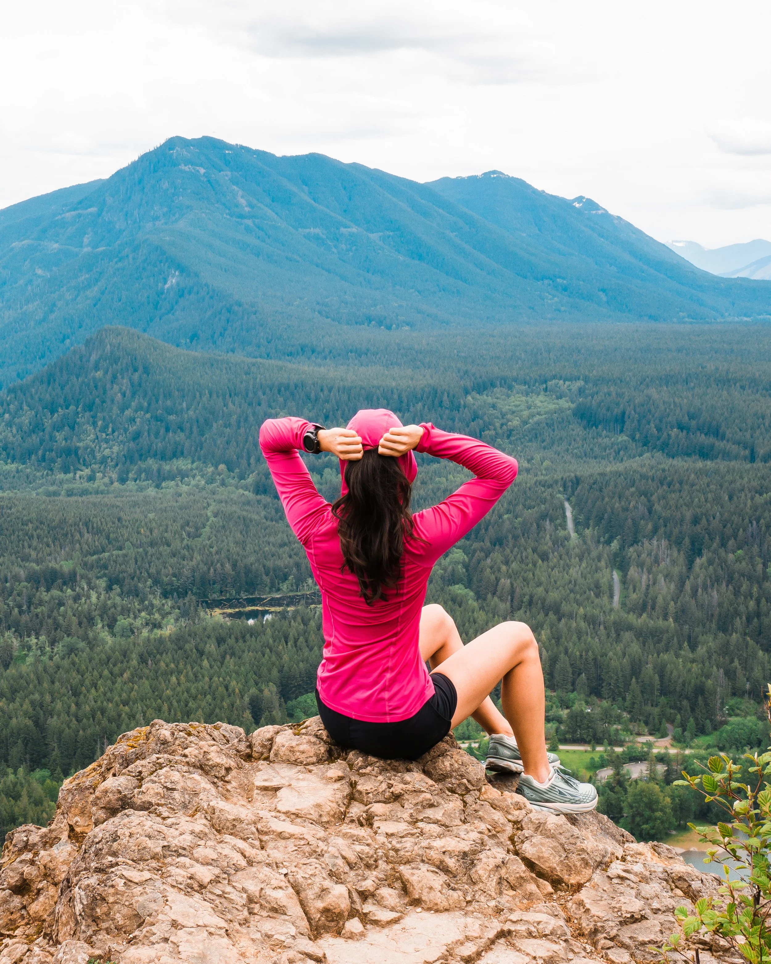 A woman in a pink long-sleeve shirt and shorts sitting on a rocky ledge overlooking a forested mountain landscape, with her hands behind her head.