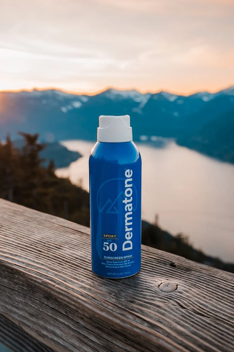 A blue bottle of Dermatone sunscreen spray with SPF 50 resting on a wooden surface outdoors during sunset, with mountains and a body of water in the background.