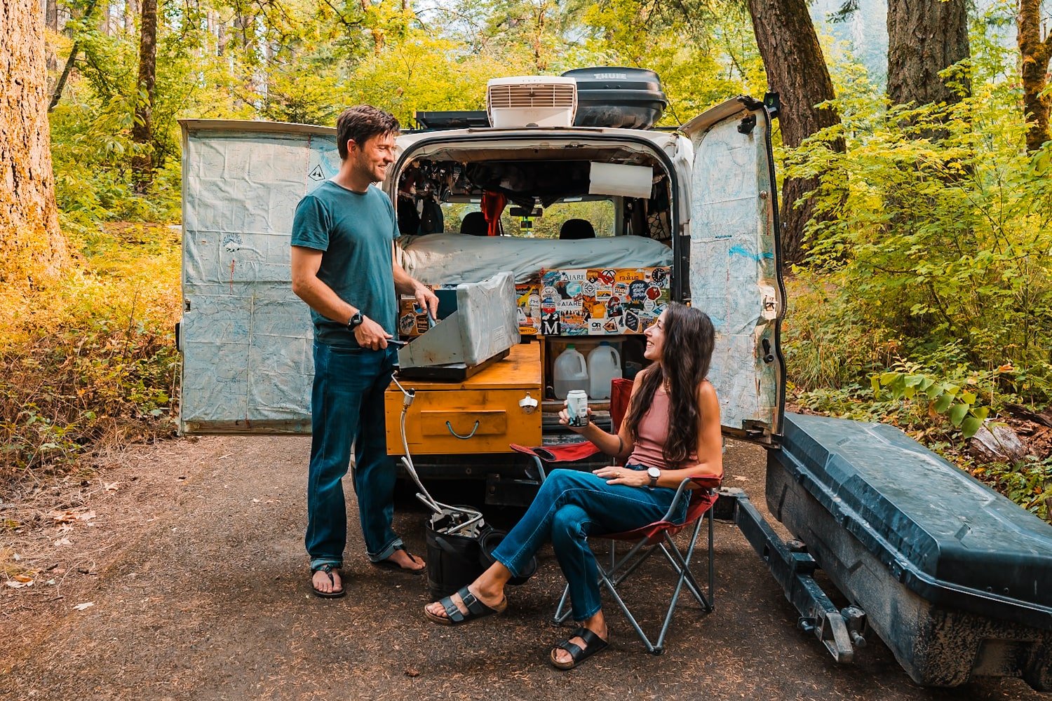 a couple at a campground in banff, next to their campervan. He is cooking on a camp stove out back and she is sitting in a camp chair.