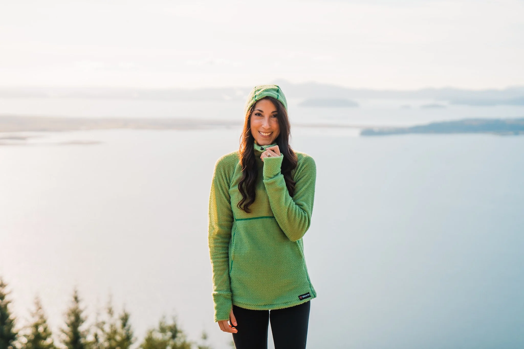 A woman wearing a light green fleece hoodie with the hood up, standing in front of water and islands