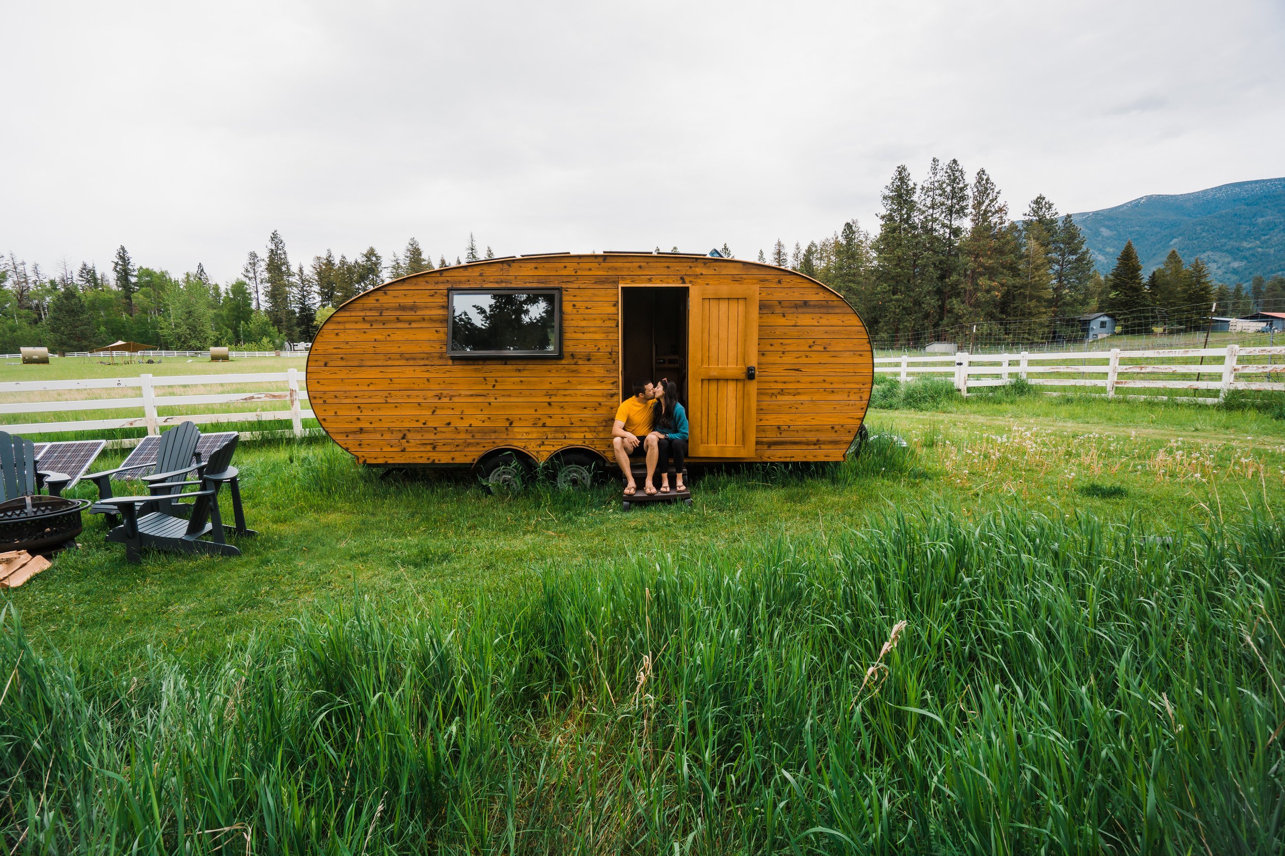 A couple sitting on the steps of a small, oval-shaped wooden camper trailer in a grassy field, with a white fence, trees, mountains, and cloudy sky in the background.