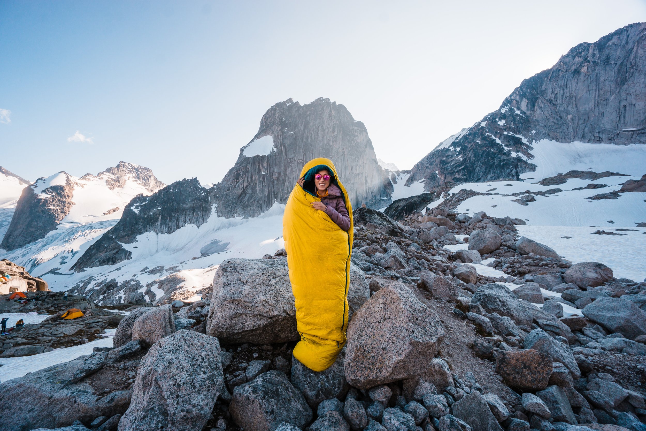 A woman wrapped in a yellow sleeping bag standing on rocky terrain in a snowy mountain landscape with peaks and glaciers in the background.