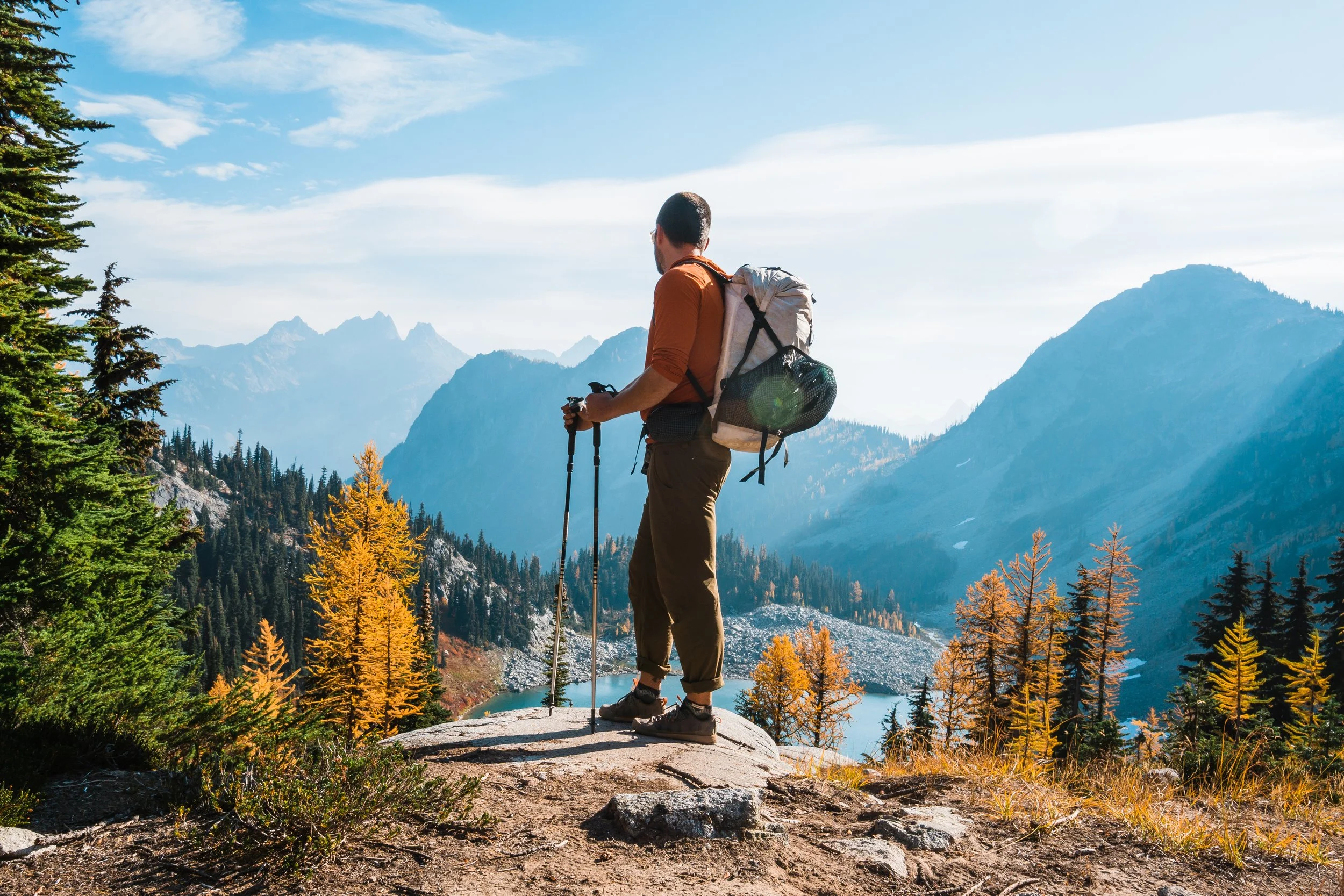 Hiker standing on a rock ledge overlooking a mountain lake surrounded by evergreen trees with autumn foliage, mountains in the background, holding trekking poles, wearing a backpack and outdoor clothing.