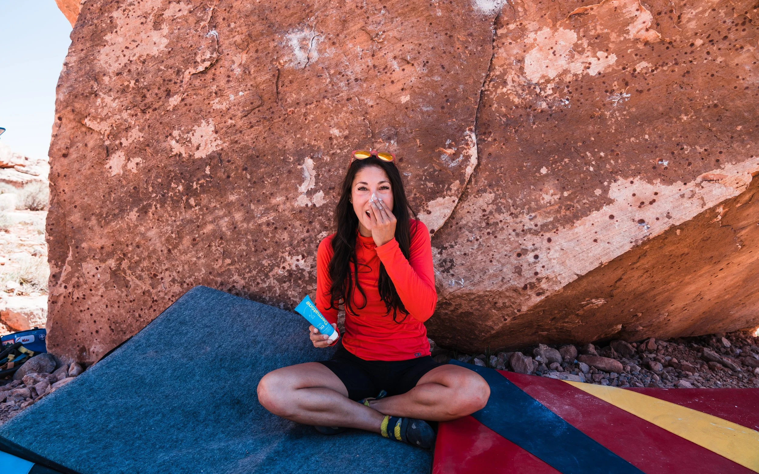 Woman with long dark hair, red long-sleeve shirt, and black shorts sitting cross-legged on a red and blue mattress against a large red rock in a desert landscape, holding a natural sunscreen tube and smiling while covering her nose.
