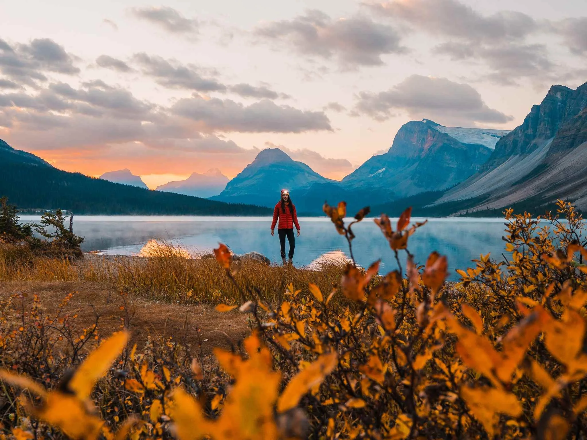 women standing by an alpine lake at sunrise wearing a red jacket and headlamp, with mountains in the background and fall colors in the foreground
