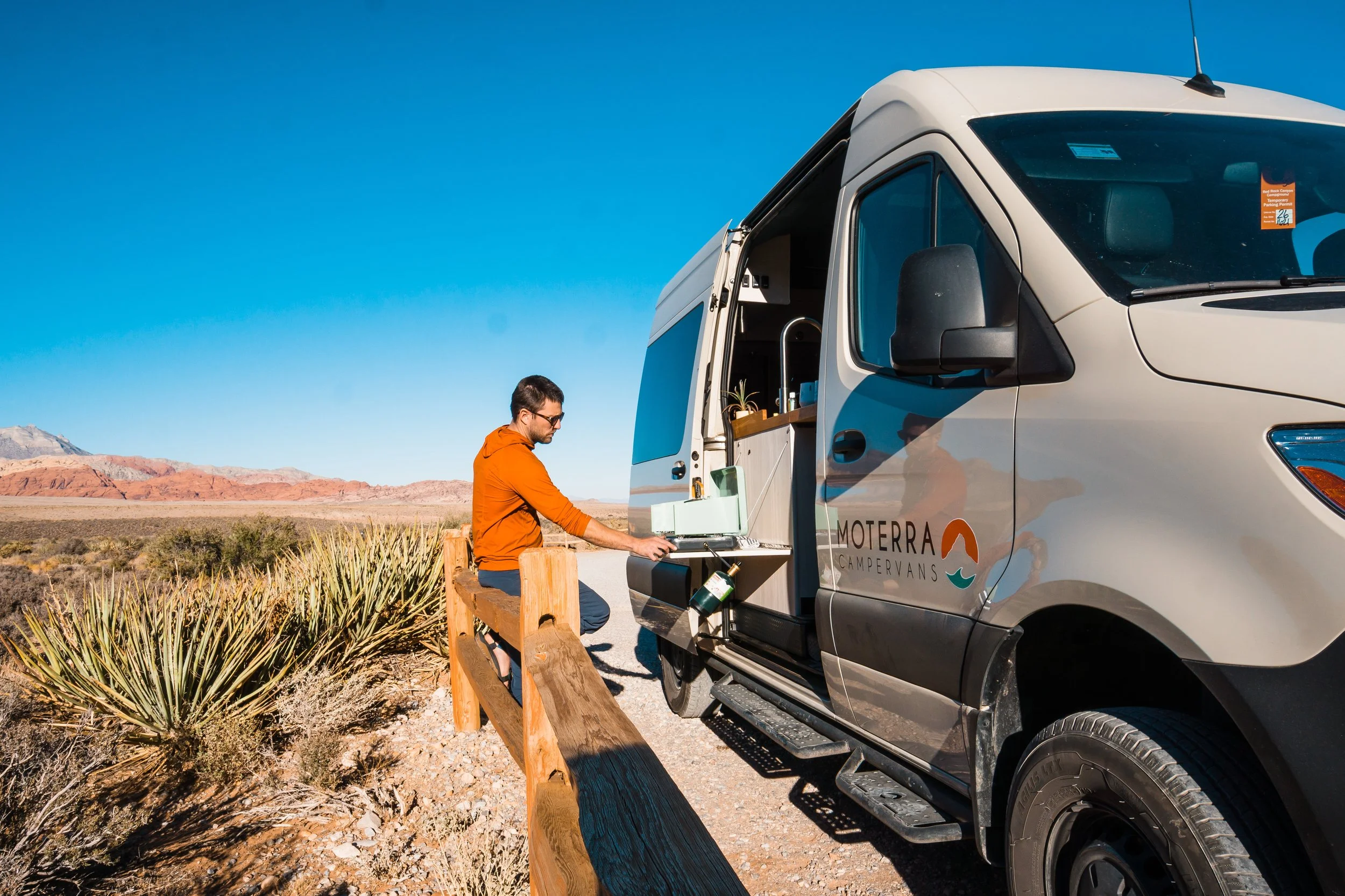 A man in an orange jacket filling a portable gas stove on the side of a camper van in a desert landscape with mountains in the background.
