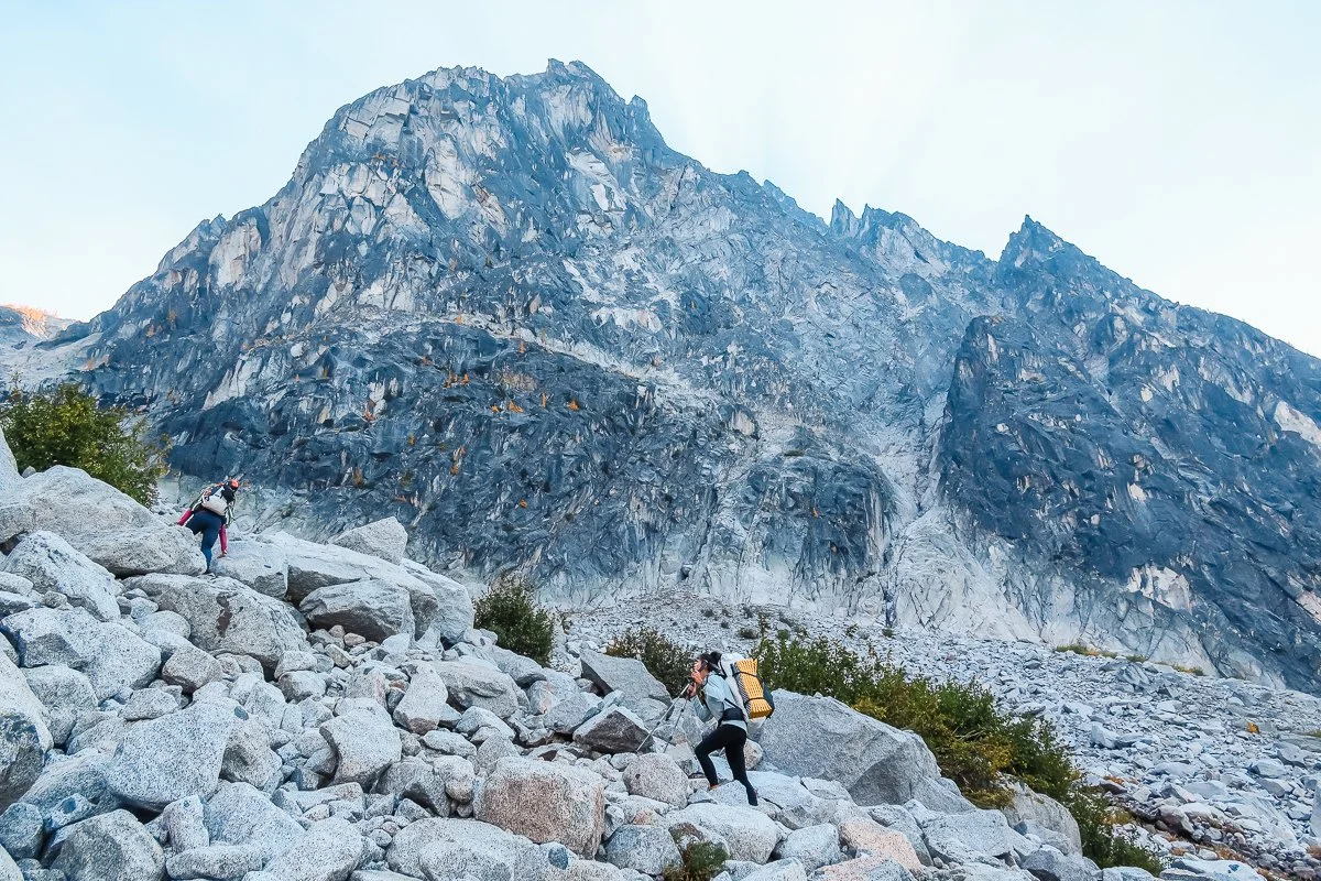 Backpackers heading up a boulder field on Aasgard Pass in the enchantments with a huge, rocky dragontail peak in the background