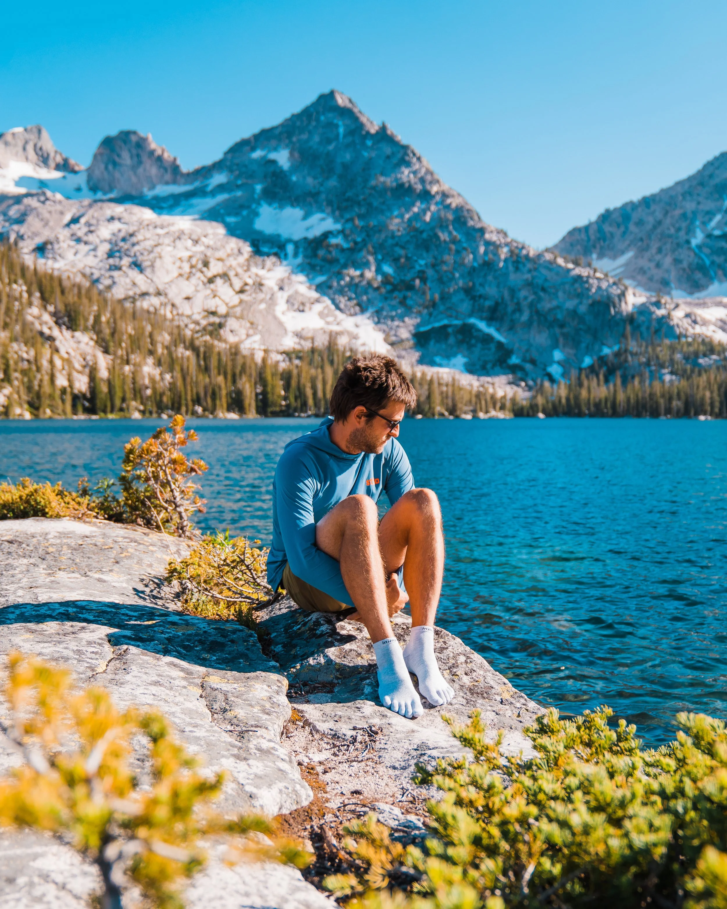 A man sitting on a large rock at the edge of a mountain lake, with snow-capped mountains and trees in the background, wearing a blue jacket and white socks.