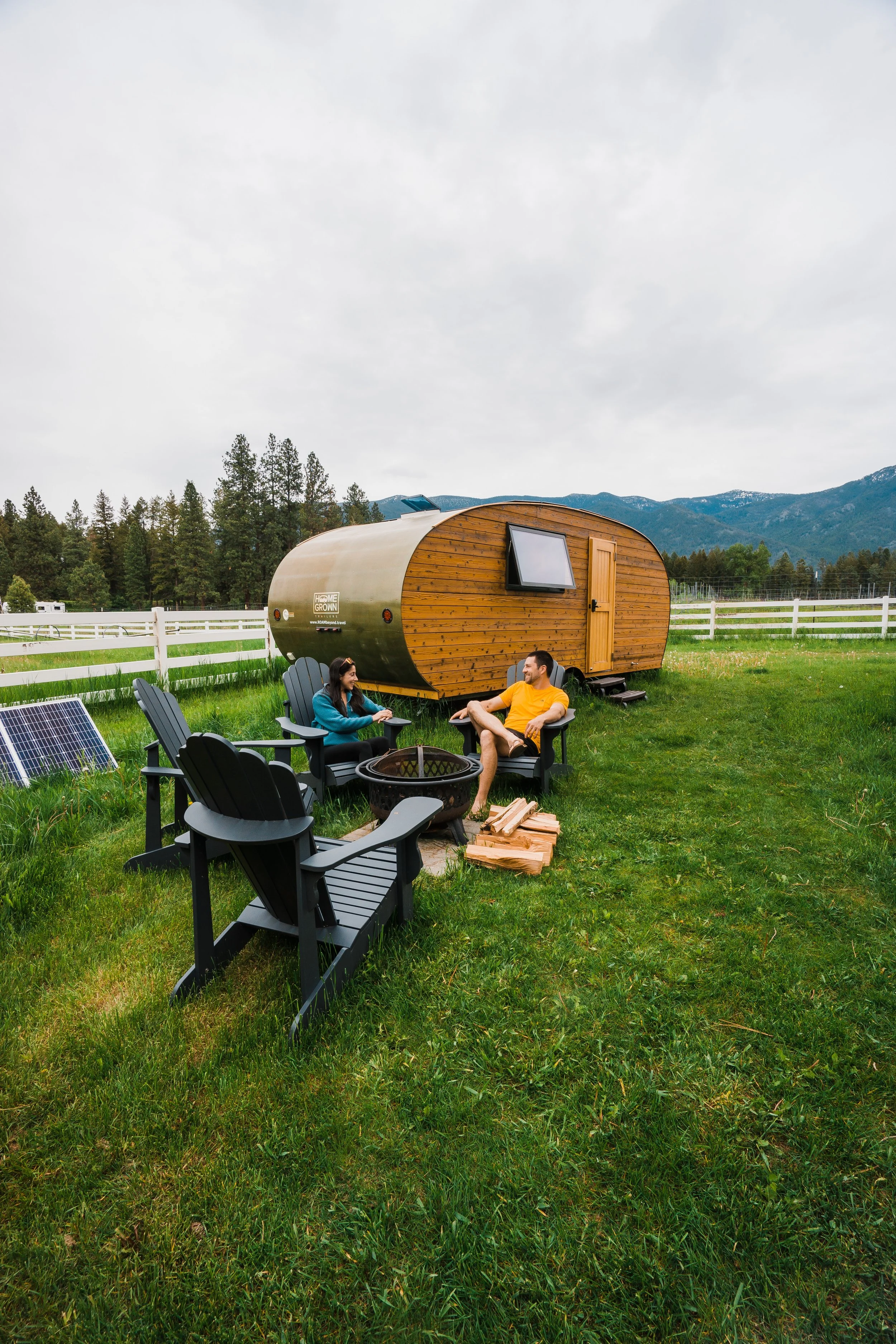 Two people sitting around a firepit outside a tiny travel trailer, surrounded by green grass and mountains in the background.