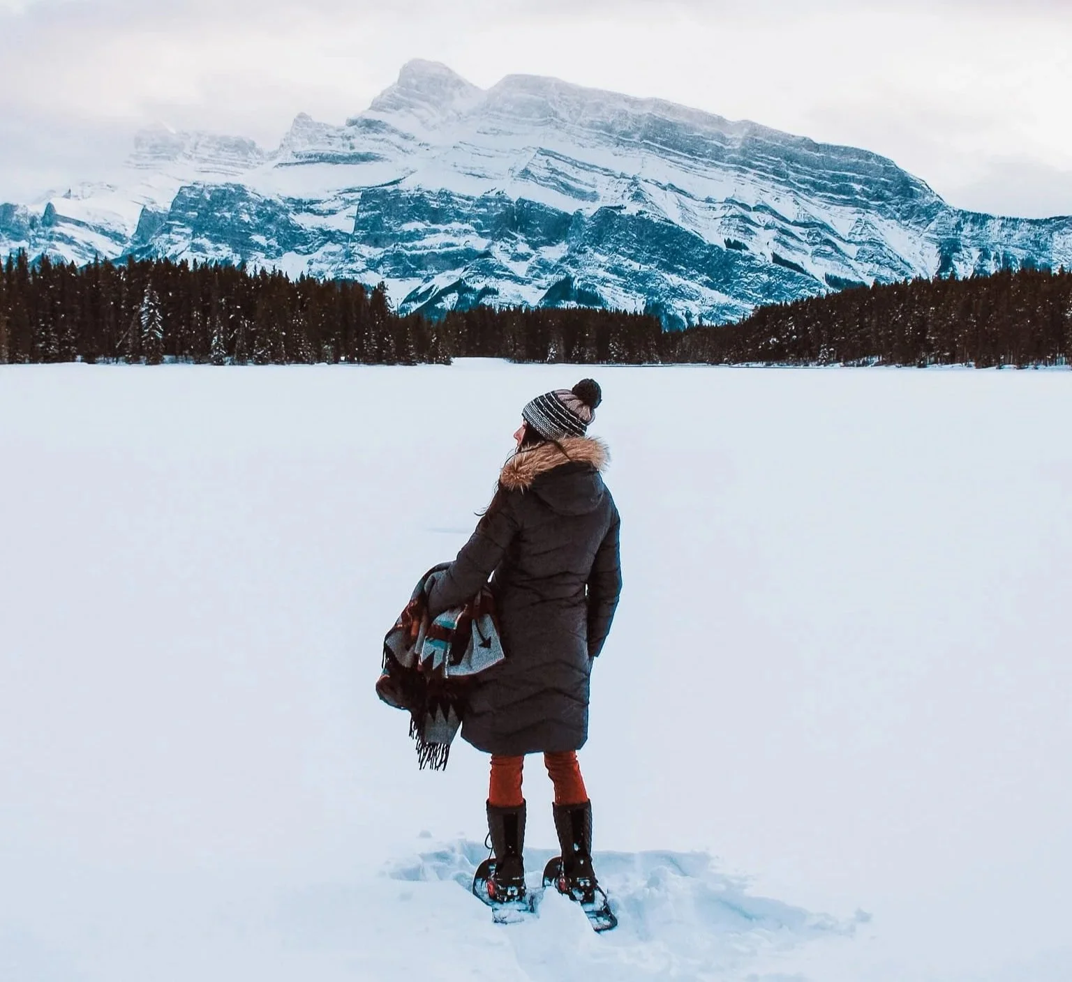 a woman standing on a snowy Two Jack Lake in banff, wearing a long down parka and snowshoes, with mount rundle in the background