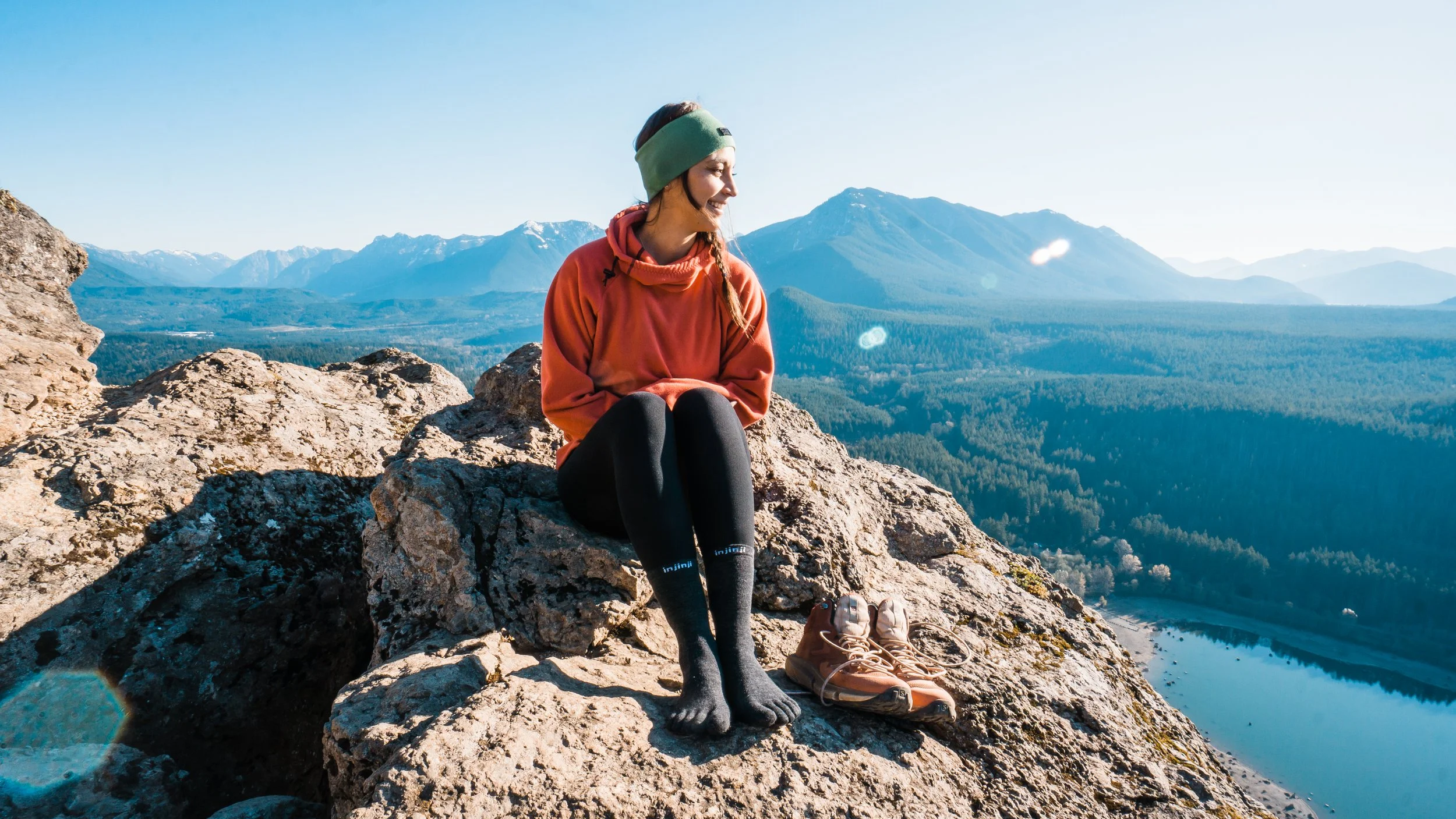 A woman sitting on a rock on a mountain peak, overlooking a forested valley and mountain range with a lake below, wearing outdoor gear and smiling.