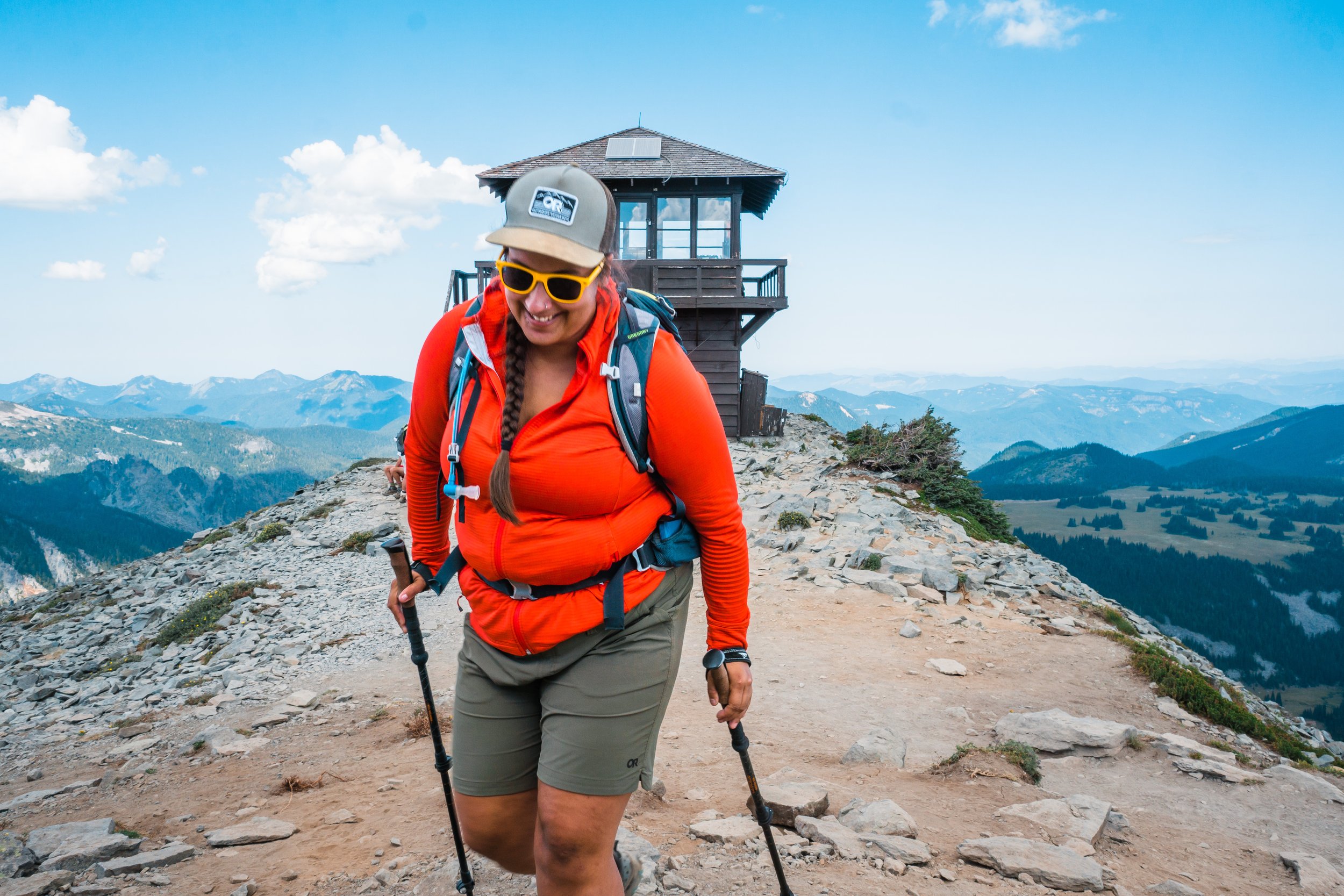 A woman hiking on a rocky mountain trail with scenic mountain range in the background. She is wearing an orange jacket, khaki shorts, yellow sunglasses, and a hat, and is carrying a backpack and trekking poles.