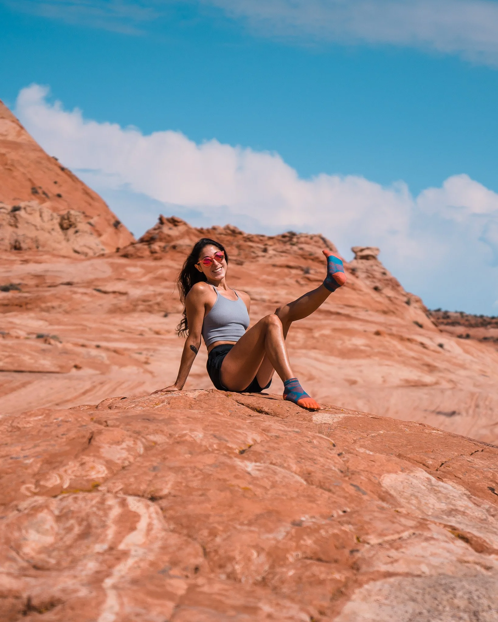 A woman wearing sunglasses, a gray tank top, black shorts, and colorful socks sits on a large red rock formation in a desert landscape with blue sky and white clouds overhead.