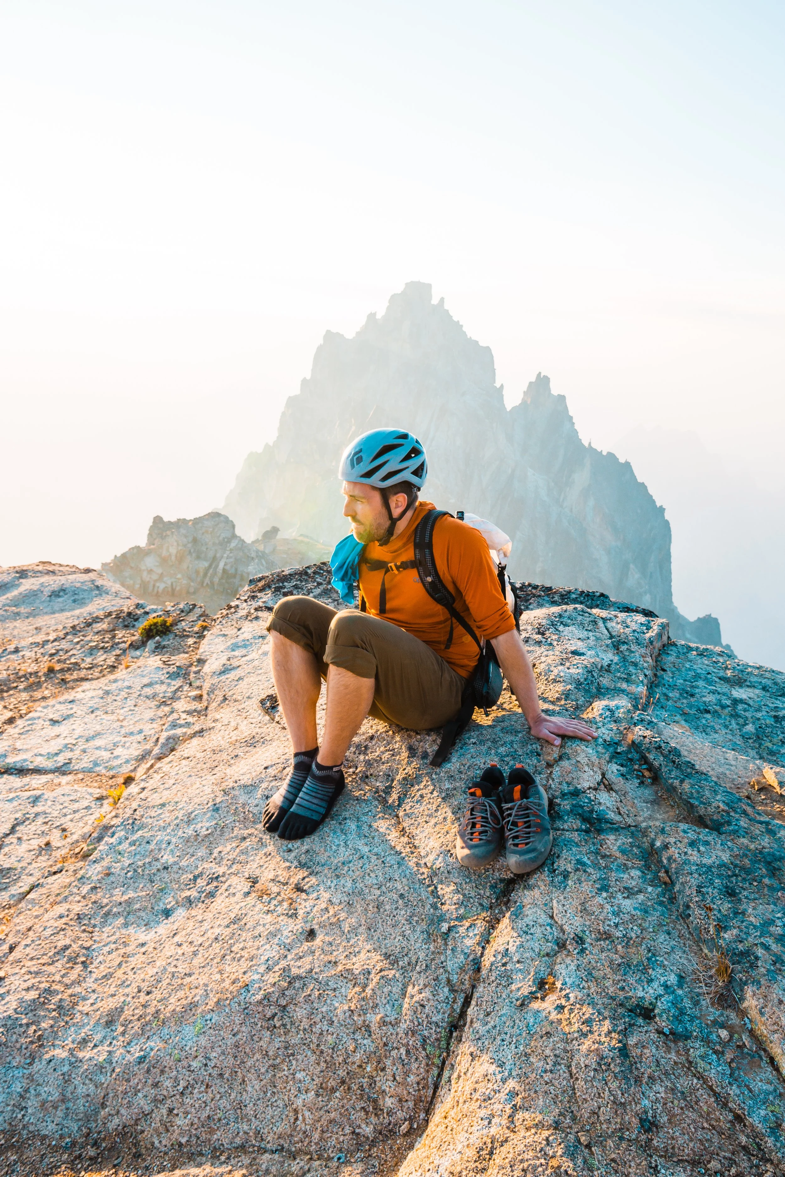 A man resting on a rocky mountaintop with a mountain peak in the background, wearing a helmet, outdoor gear, and hiking shoes placed beside him.