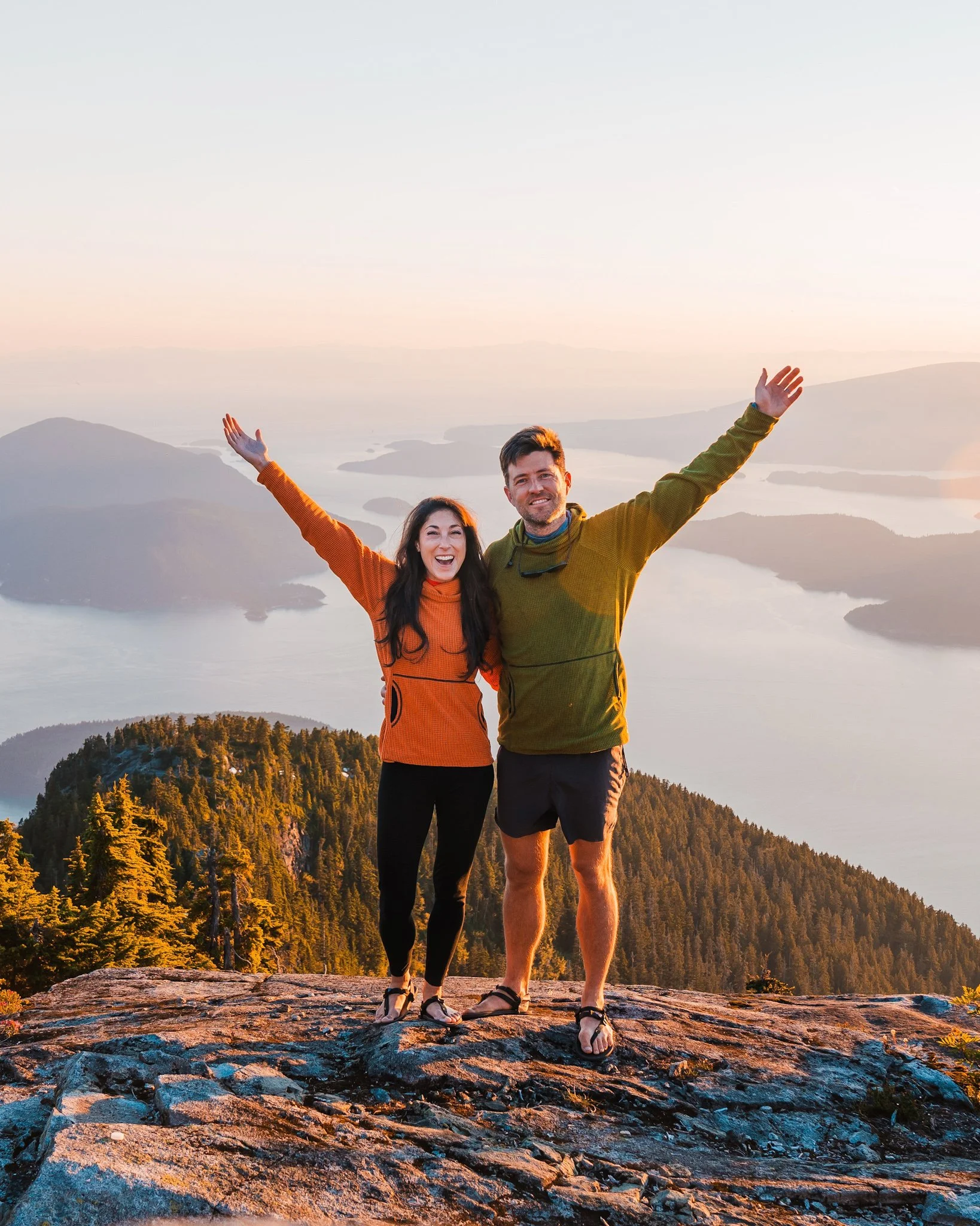 a couple smiling for the camera at sunset, with their arms raised, in front ov a view of the Howe Sound in British Columbia
