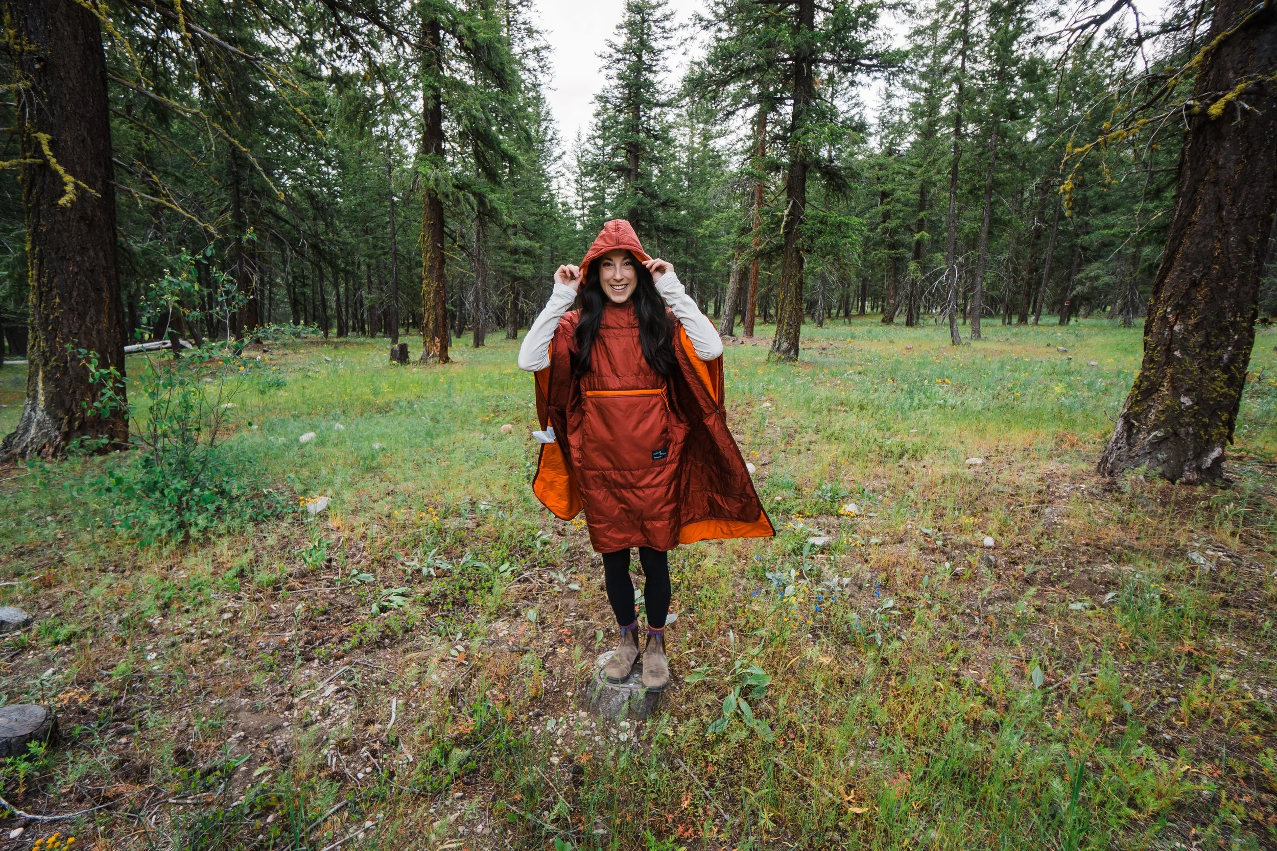 A woman in a red raincoat and backpack smiling while standing on a small tree stump in a forest.
